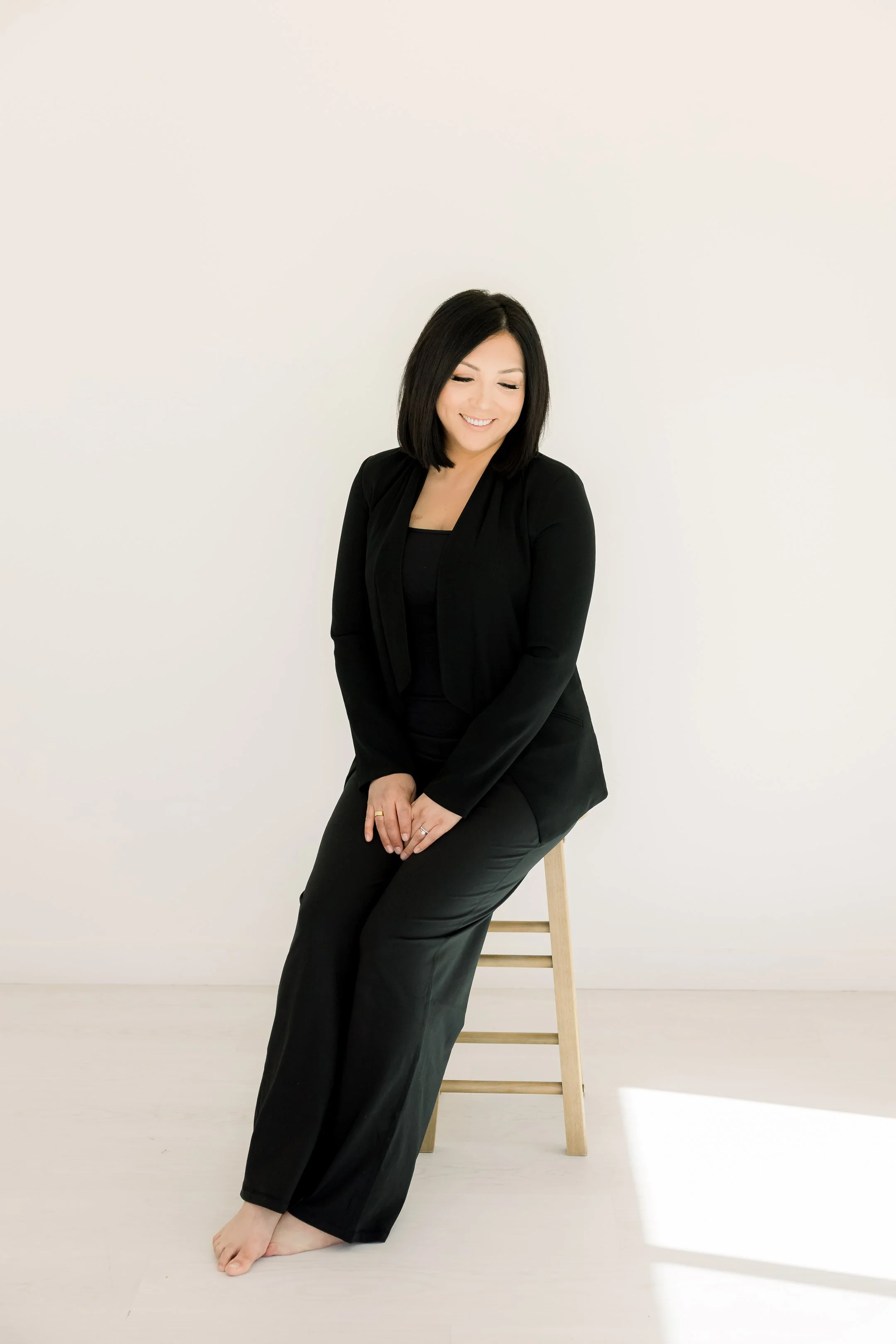 A woman in black clothing sitting on a wooden stool against a plain white background, smiling and looking down.
