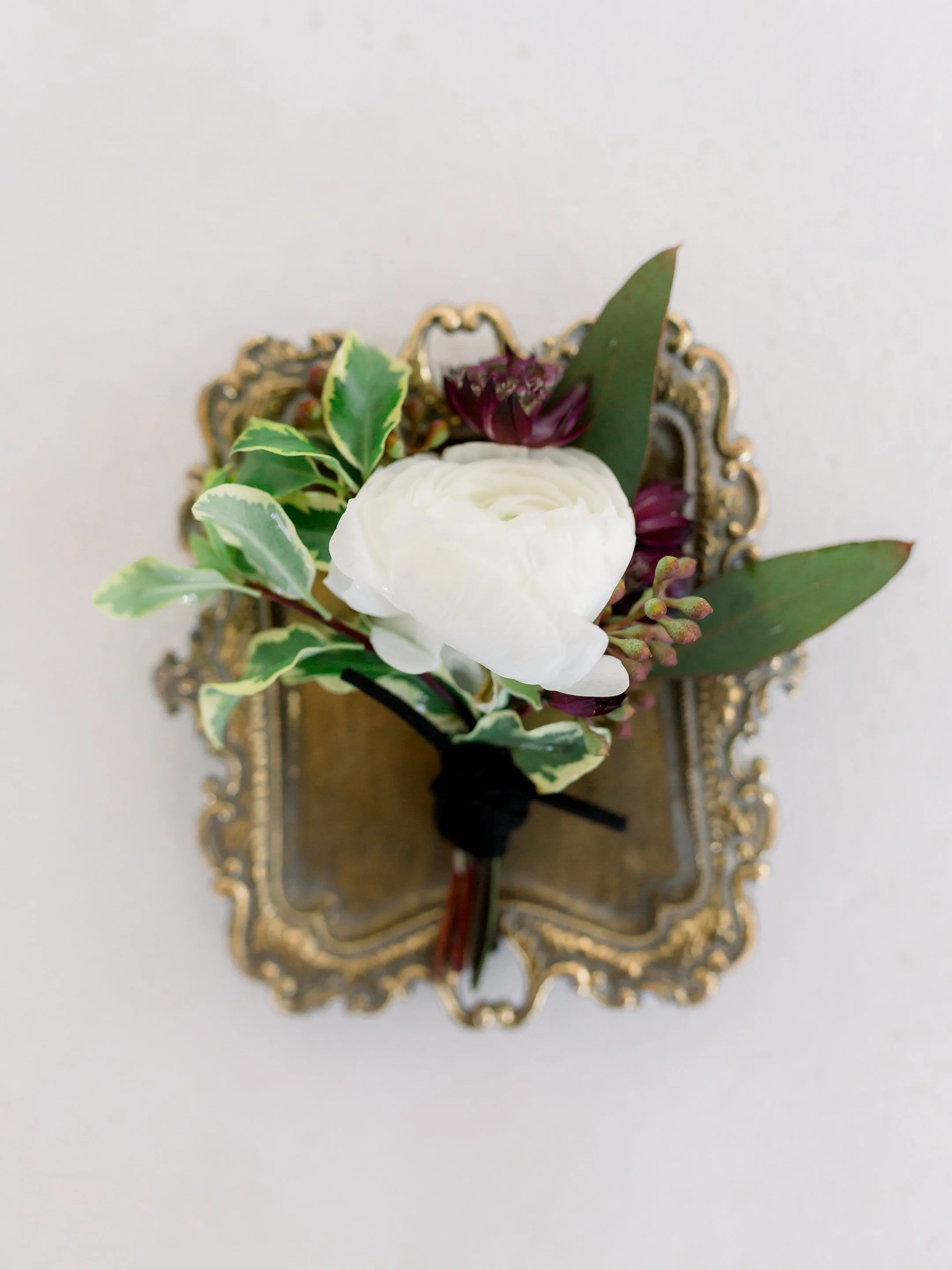 A floral boutonniere with a white ranunculus, purple flowers, and green foliage, attached to a decorative gold frame background.