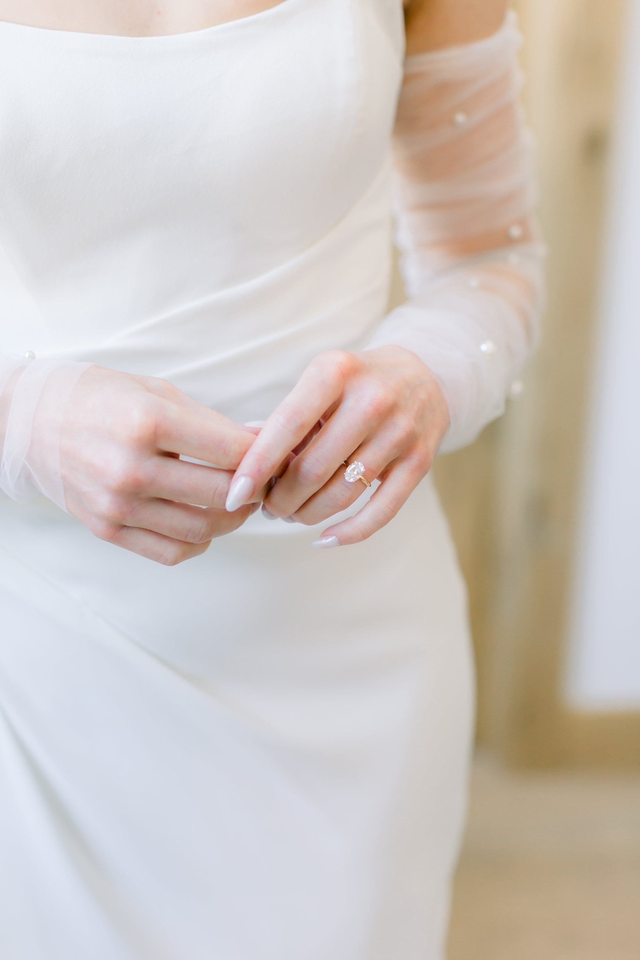 A bride wearing a white wedding gown with sheer sleeves adorned with small pearls, holding a diamond engagement ring on her ring finger.