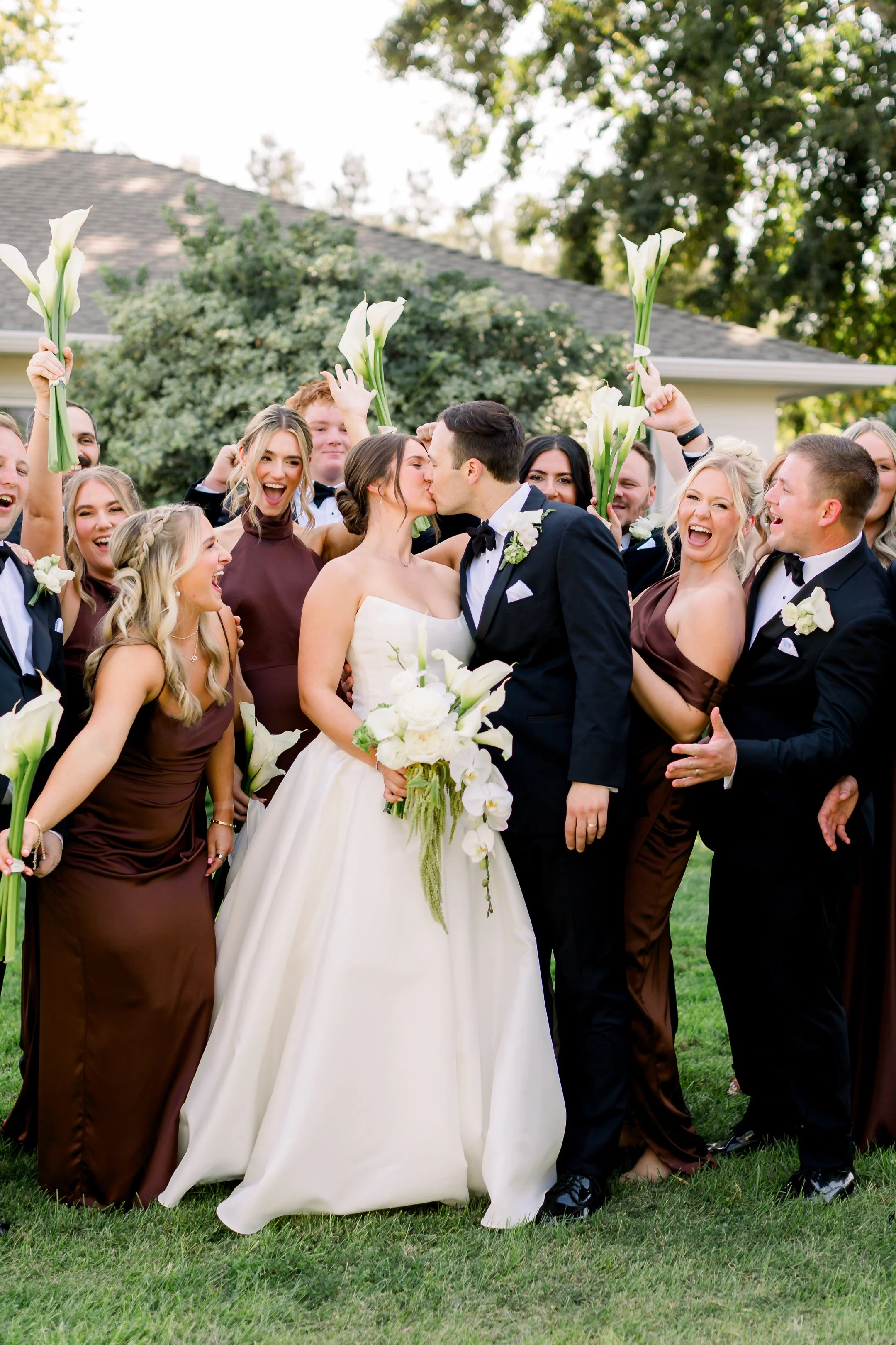 A wedding celebration with the bride and groom kissing surrounded by friends and family holding calla lilies in an outdoor setting.