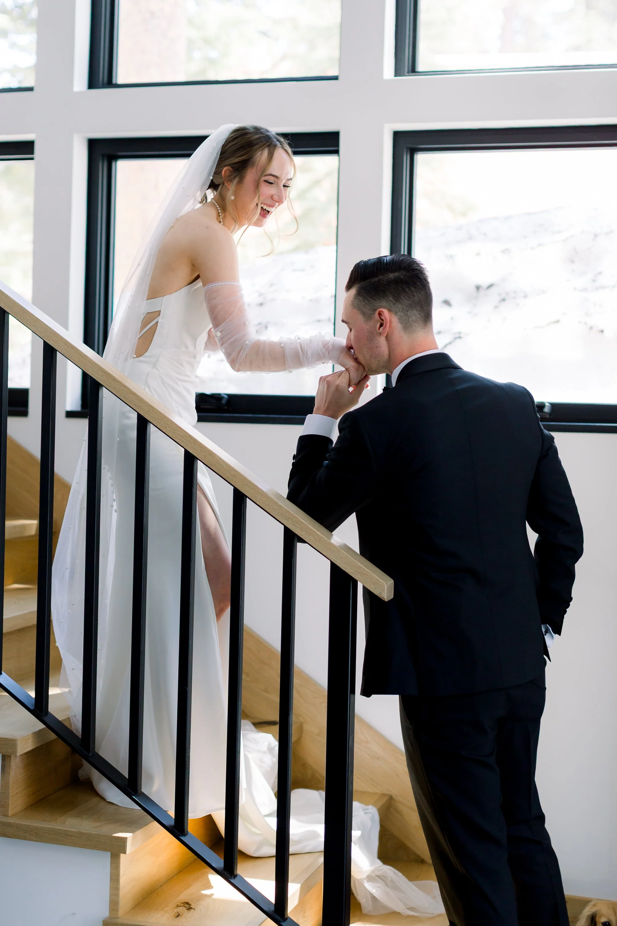 A bride and groom on a staircase, with the groom kneeling and kissing the bride's hand, inside a modern house with large windows.