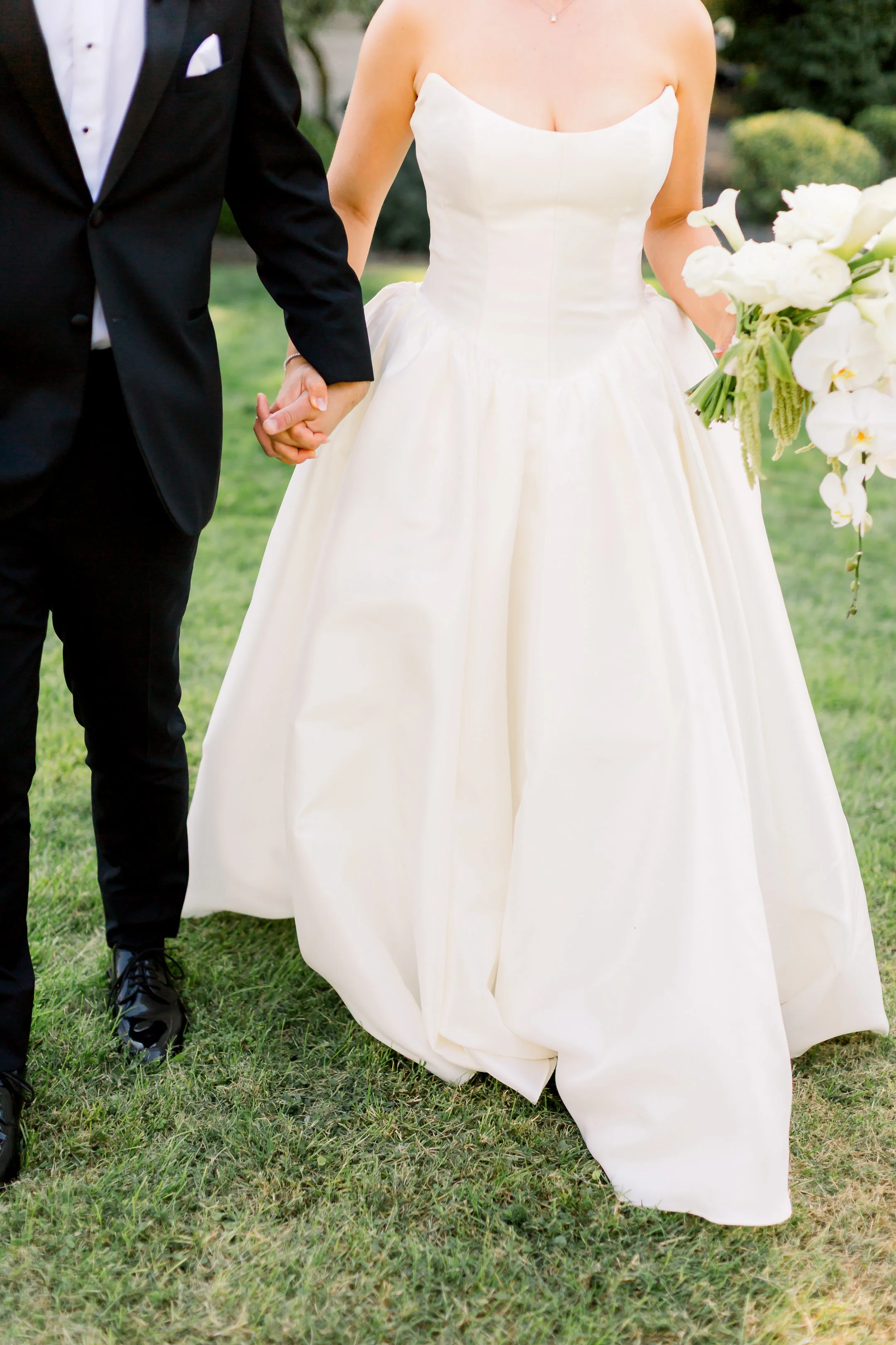 Bride and groom holding hands at their wedding outdoors, bride in a strapless white gown holding a bouquet of flowers, groom in a black tuxedo.