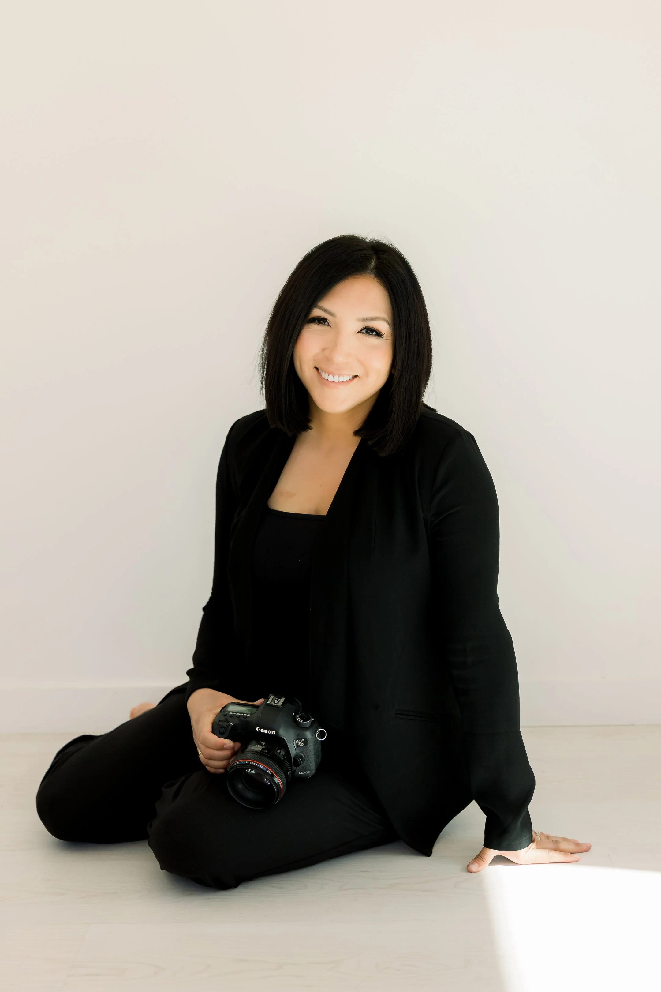 A woman with black hair, wearing a black blazer and black pants, sitting on a light-colored wooden floor, holding a professional Canon camera, smiling at the camera, against a plain white background.