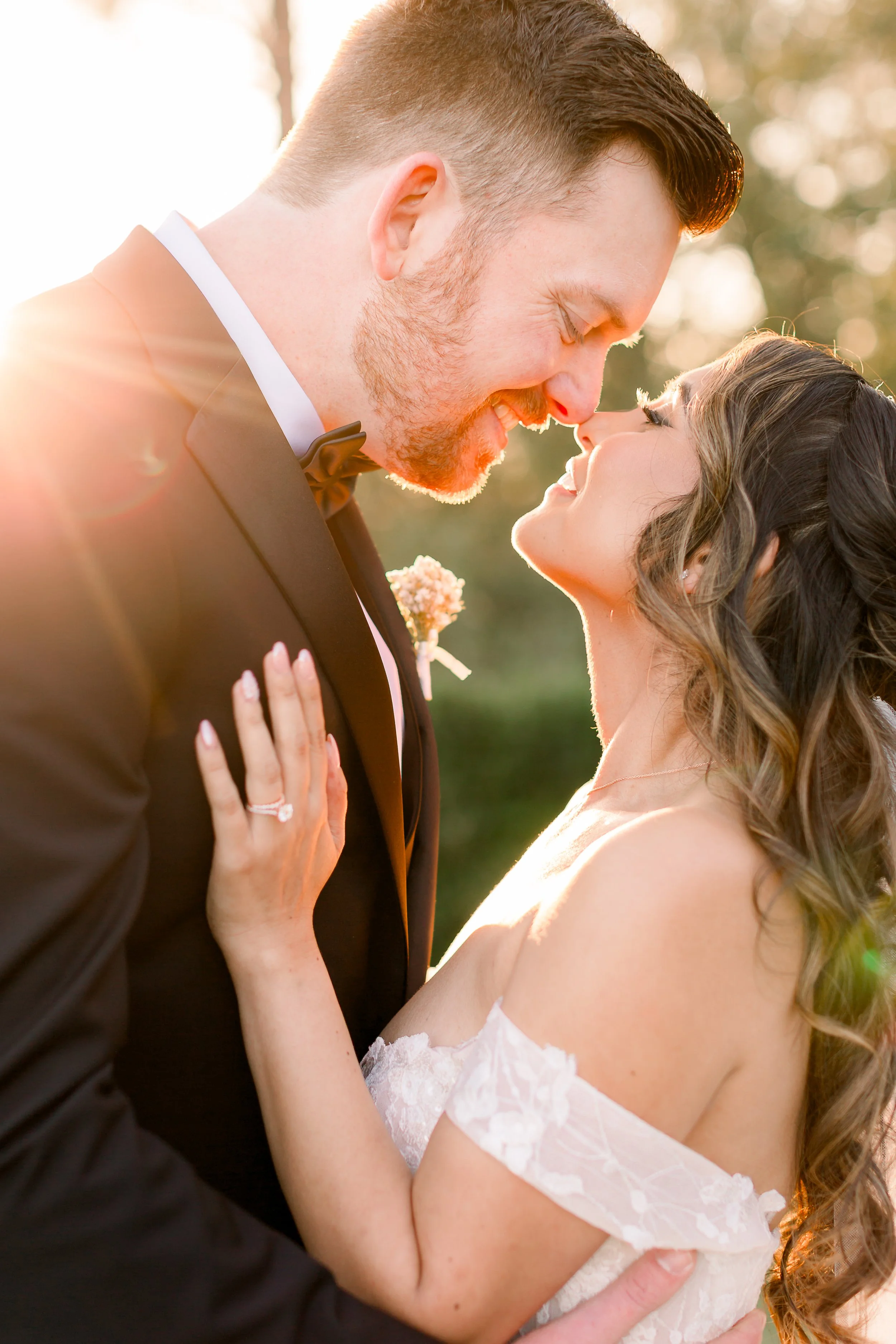 A newlywed couple sharing a romantic moment outdoors during sunset, with the groom in a black tuxedo and the bride in a white lace off-shoulder wedding dress.