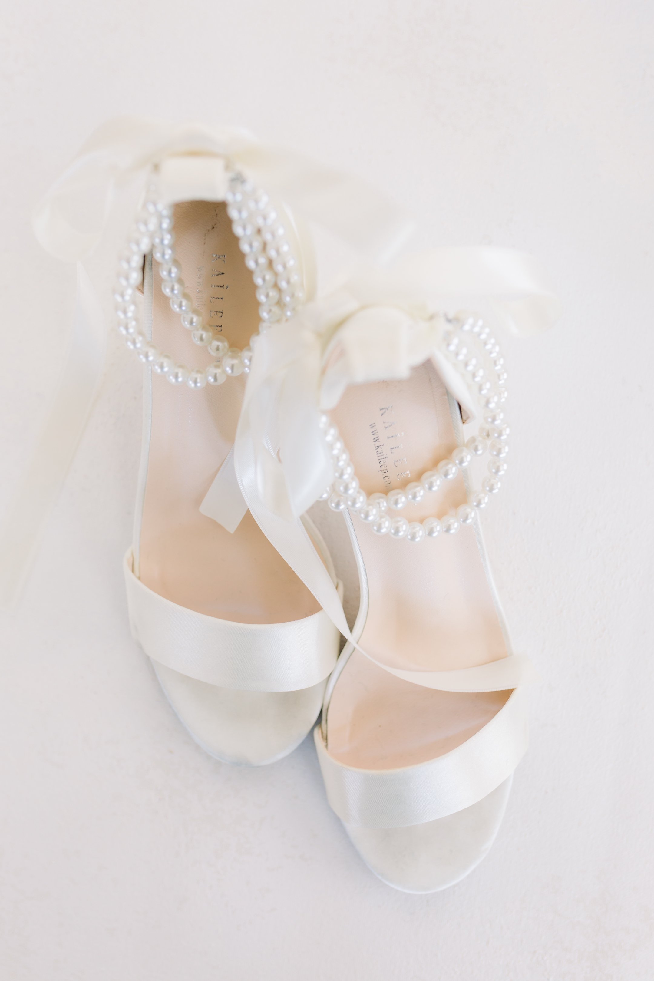 A pair of white satin bridal shoes decorated with pearl ankle straps, ribbons, and bows.