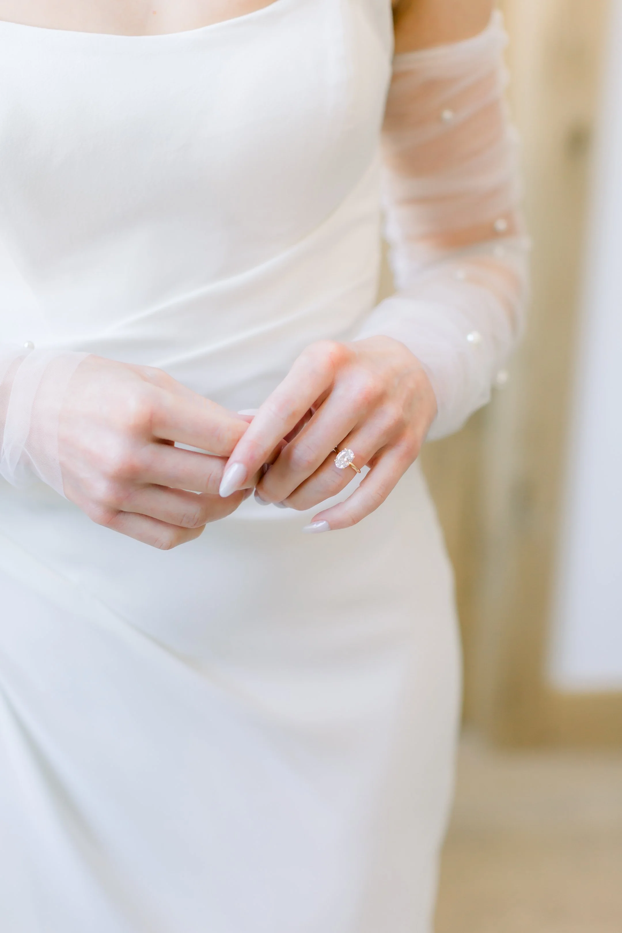 Close-up of a bride's hands adjusting an engagement ring with a large round diamond on her finger, wearing a white wedding dress with sheer, pearl-embellished sleeves.