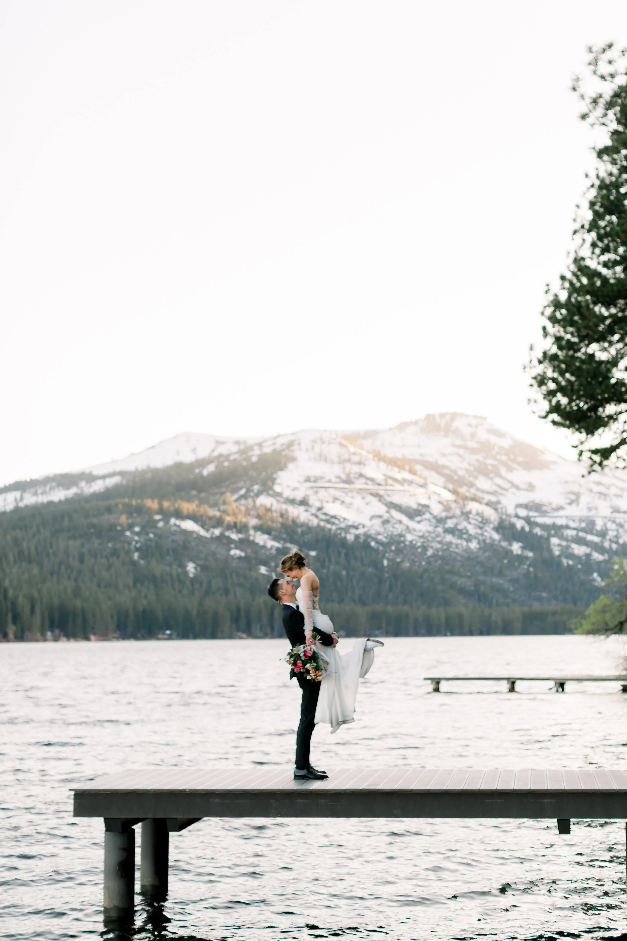 A bride and groom standing on a dock by a lake, with snow-capped mountains in the background, as the groom lifts the bride in his arms during a wedding photoshoot.