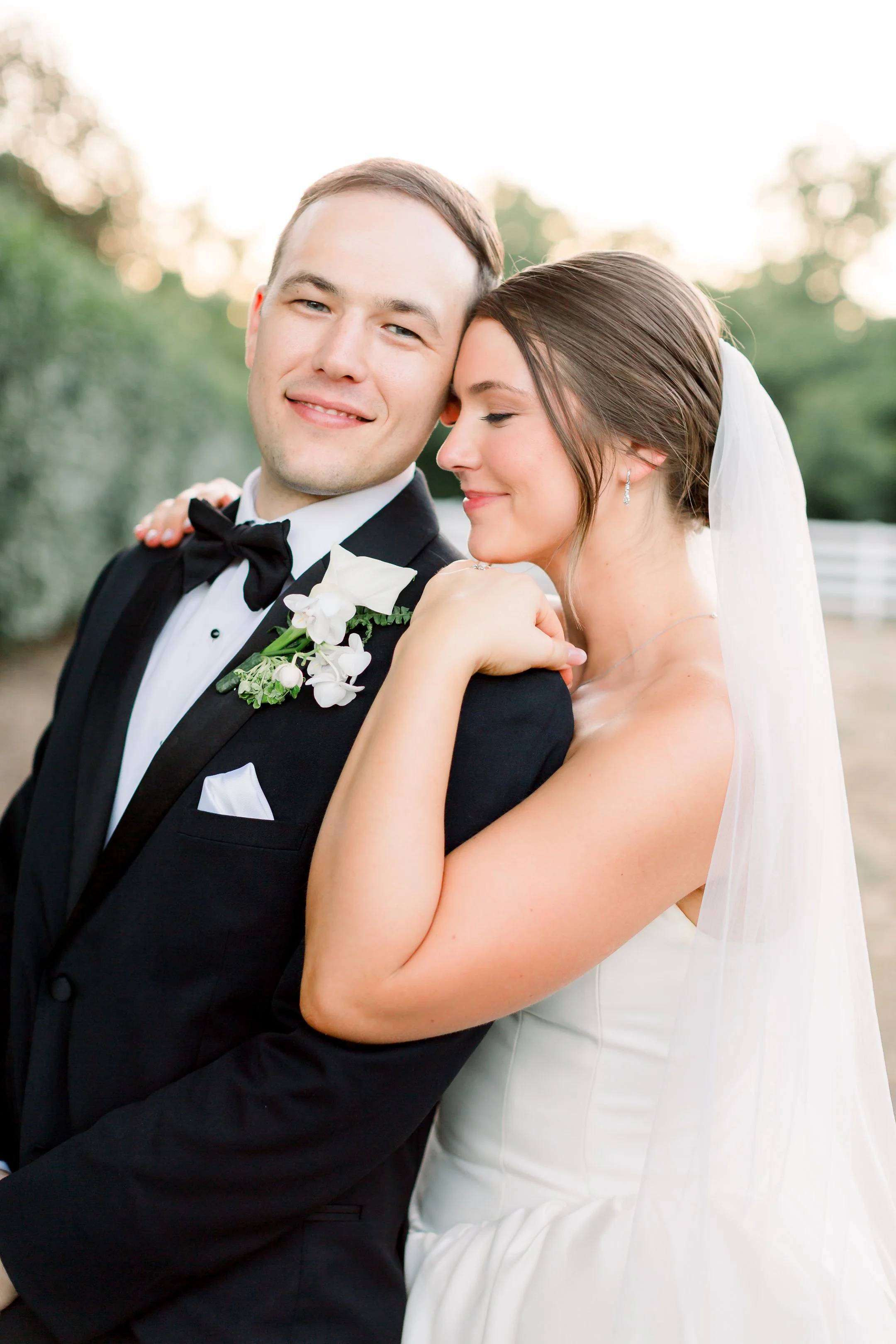 A bride and groom sharing an intimate moment outdoors, with the groom wearing a black tuxedo and the bride in a white wedding gown and veil, with greenery and a white fence in the background.