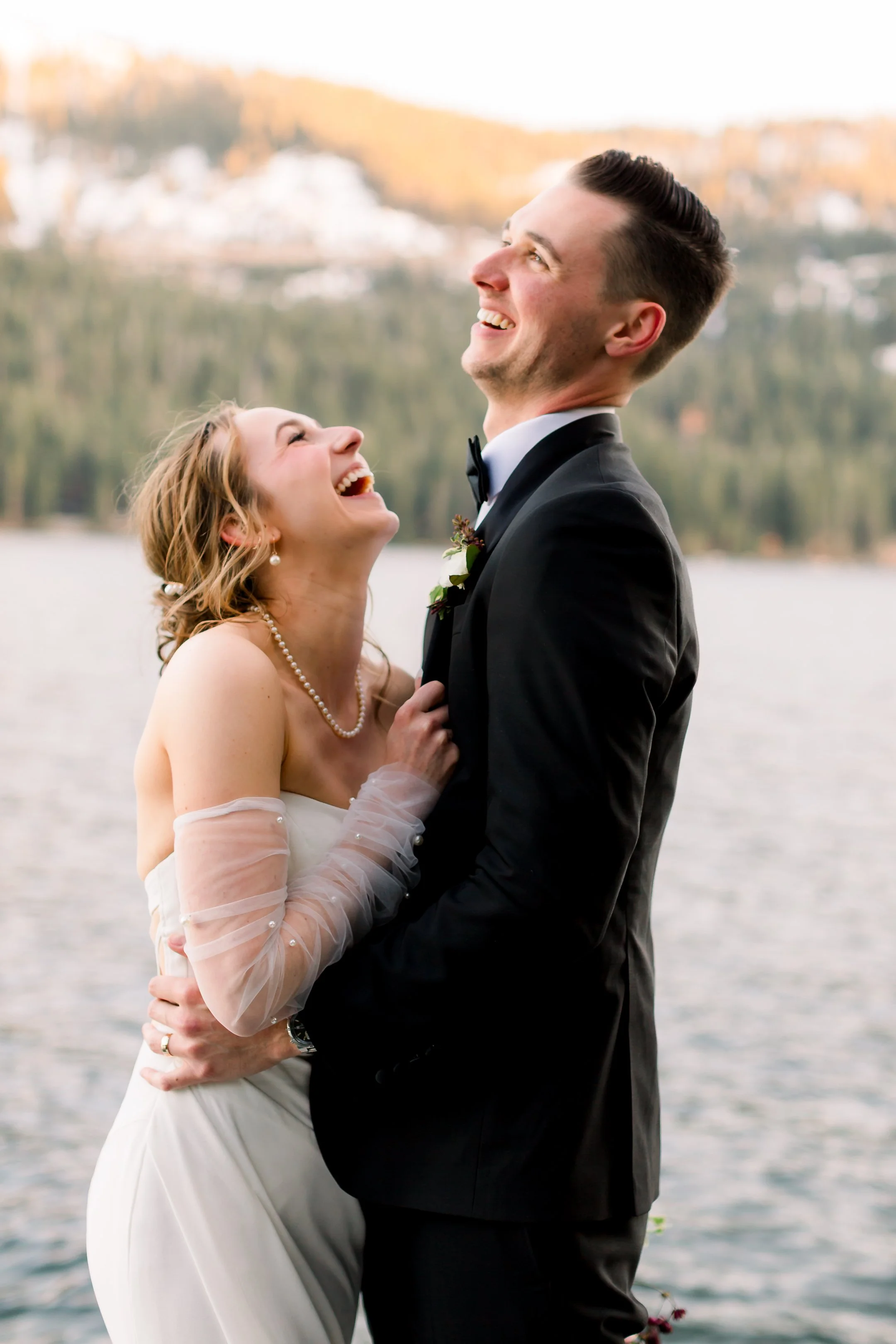 A bride and groom dressed in wedding attire outdoors by a lake, laughing and enjoying their moment.