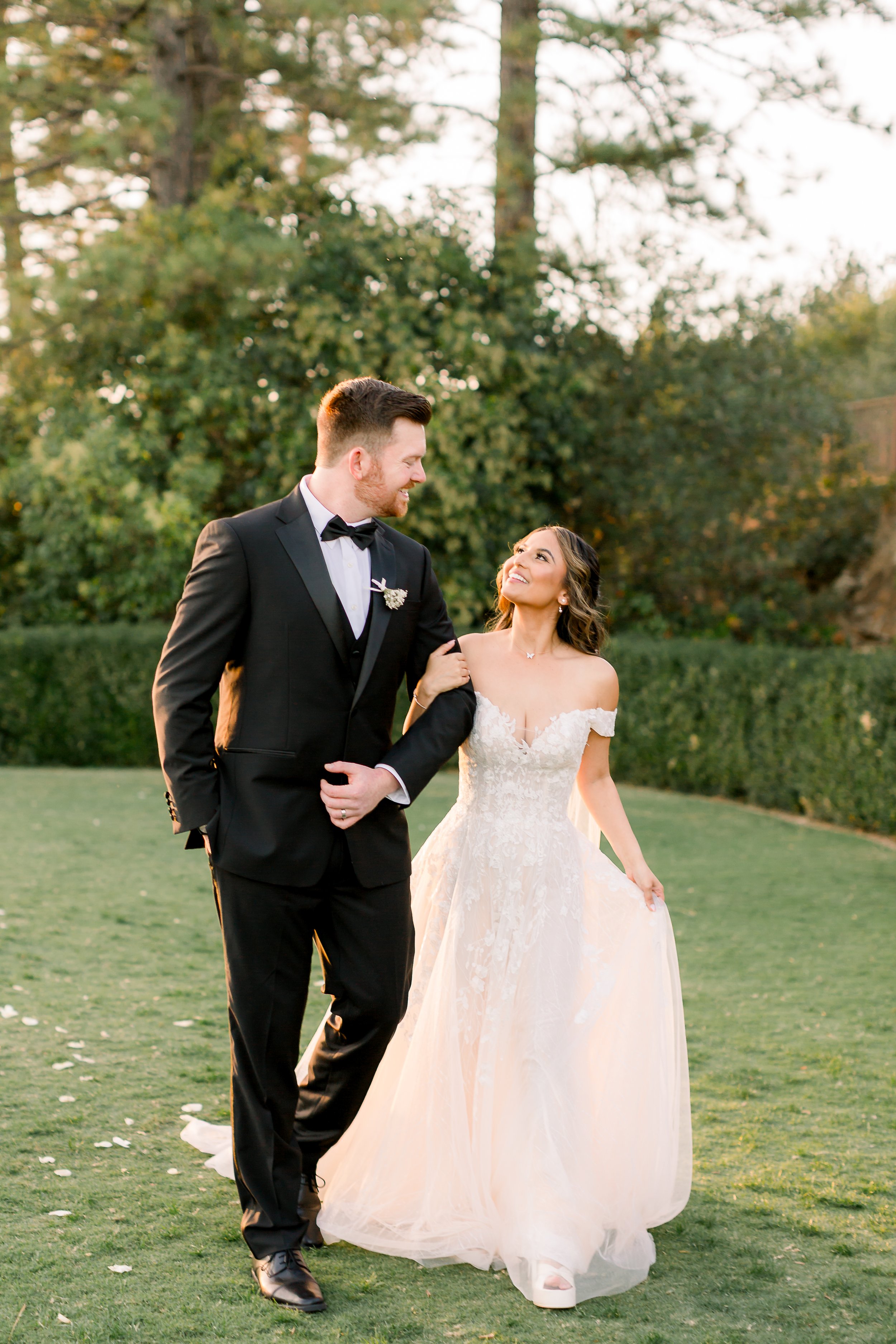 A bride and groom walking together outdoors on a grassy area during sunset, smiling at each other. The groom is wearing a black tuxedo and the bride is in a white wedding dress.