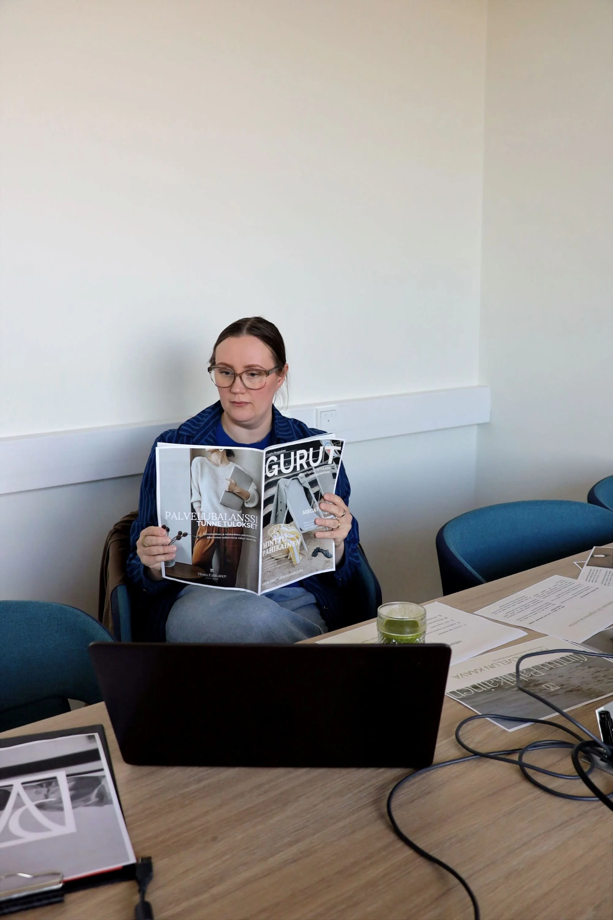 Nainen lukee lehteä kokoustilassa. Silmälasit, ruskeat hiukset, sininen Marimekon Jokapoika-paita. Lehti on kuvitteellinen asiantuntijayrittäjien julkaisu nimeltä Gurut. Pöydällä vihersmoothie kristallilasissa, läppäri ja papereita sikin sokin.