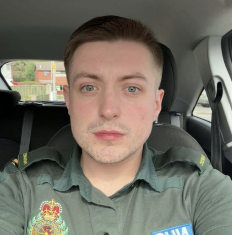 A young man with short brown hair and light skin sitting in a police uniform inside a car.