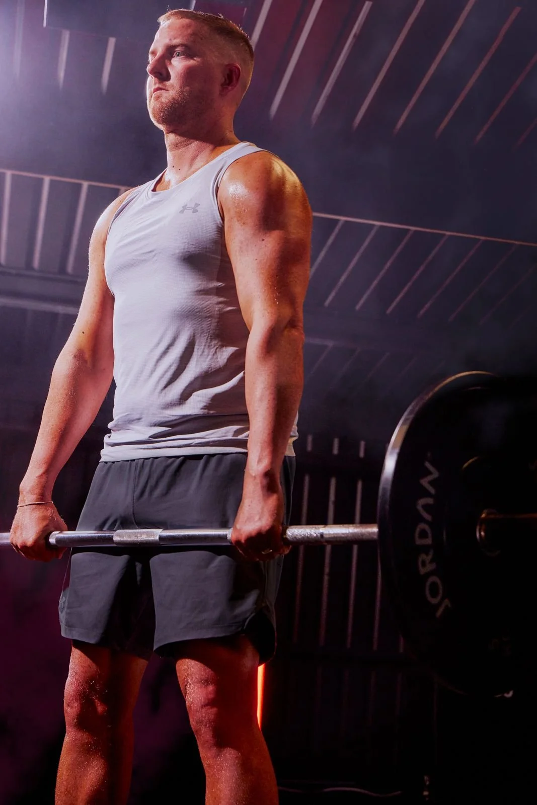 A man lifting a barbell with a focused expression, muscular arms, and sweat on his skin, in a gym setting with dark ceiling beams and colored lighting.