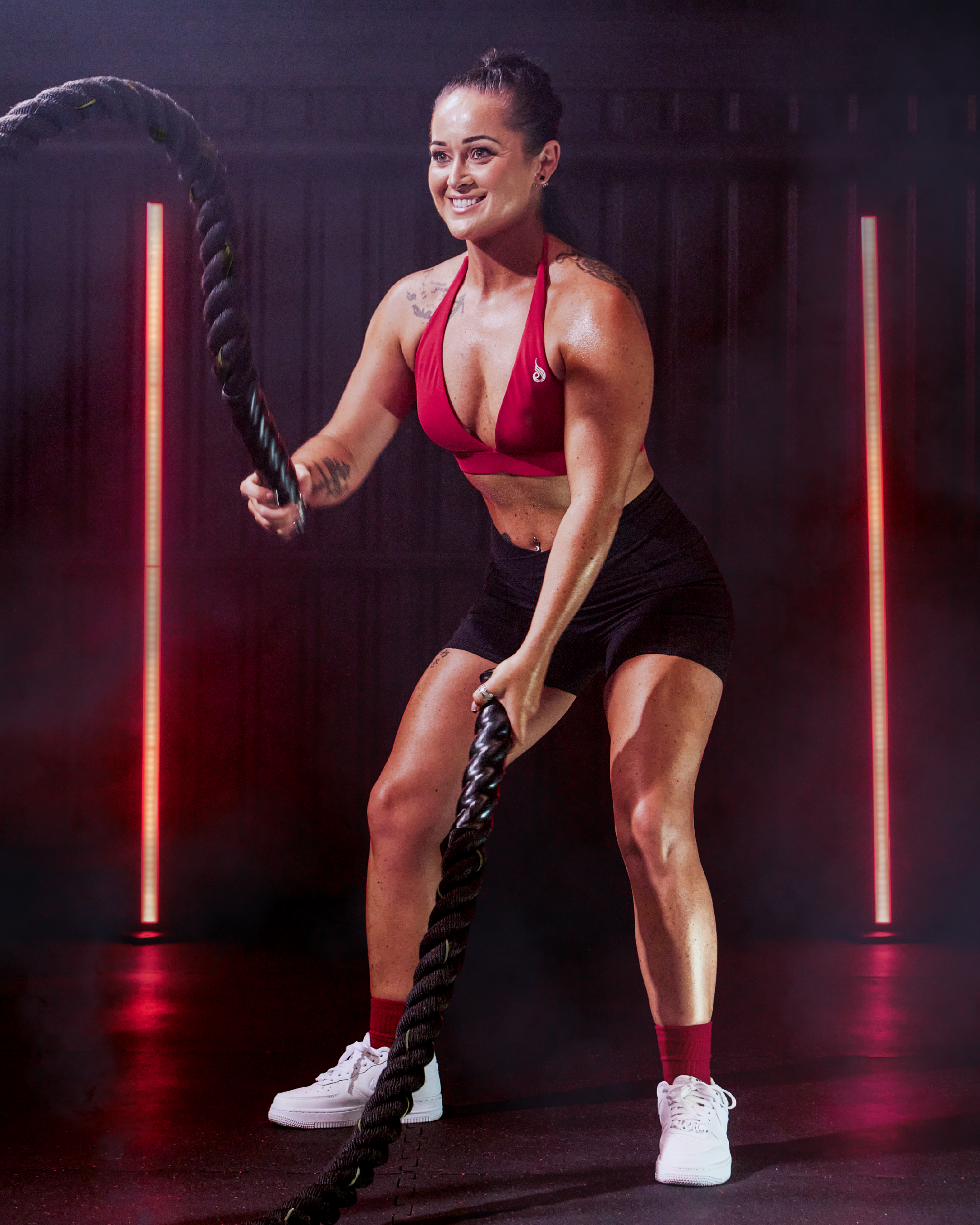 A woman with a toned physique, wearing a red sports bra and black shorts, is using battle ropes in a gym with red lighting.