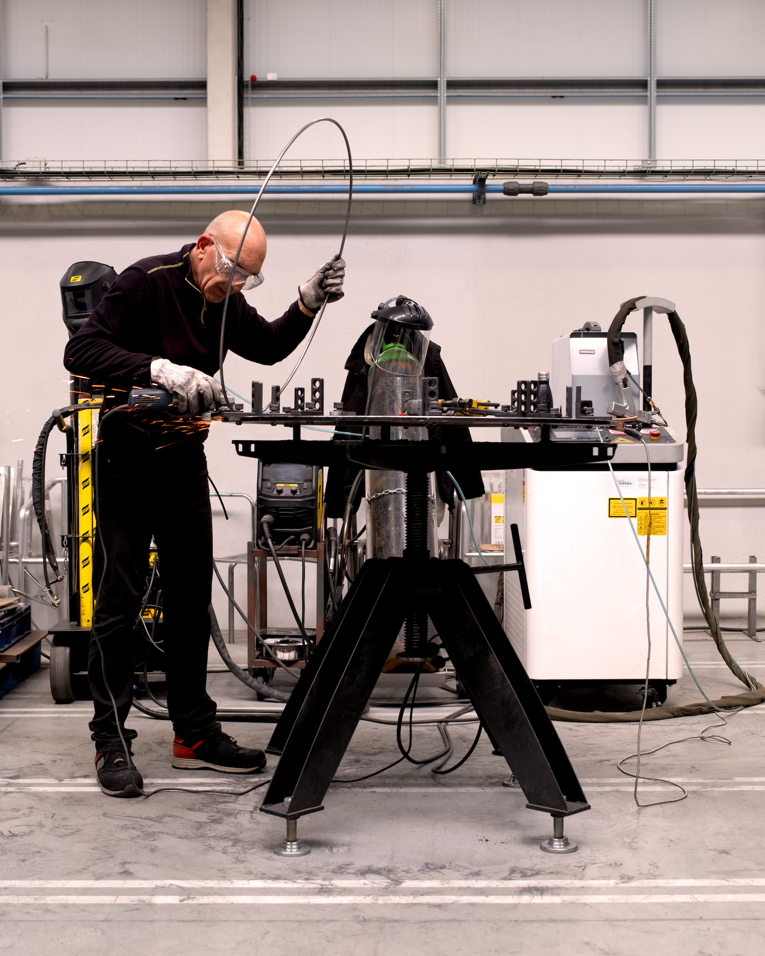 Un ingénieur effectue des travaux de soudage ou de fabrication dans un atelier industriel, utilisant un casque de protection et des gants, avec des étincelles visibles.