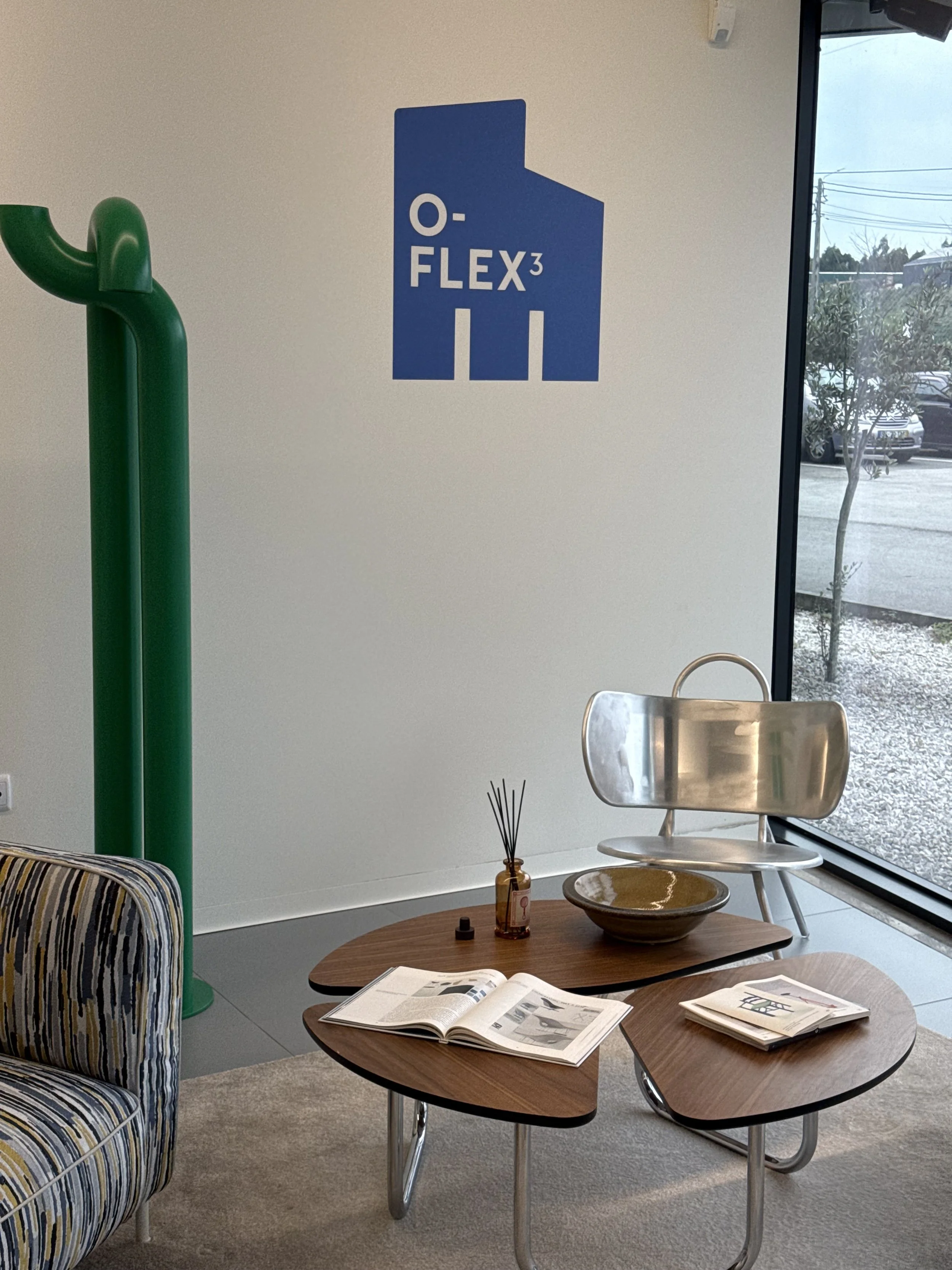 Intérieur d'un salon avec une table en bois, une chaise en métal, une cheminée en verre, un fauteuil rembourré à rayures, et une grande fenêtre avec vue sur une rue avec des voitures et des arbres, et un mur avec un logo bleu 'O-FLEX3'.