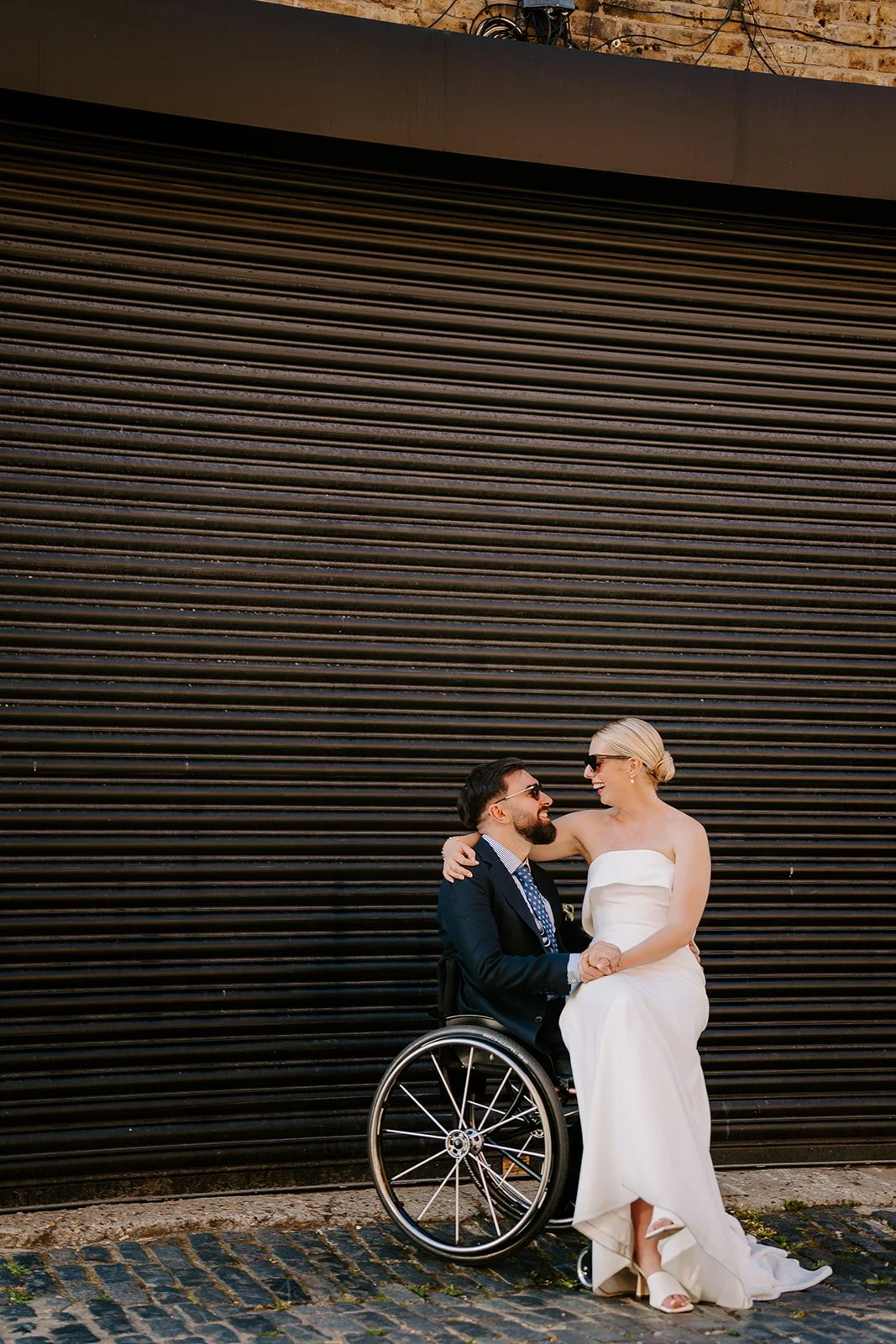 A bride sits laughing on her groom's lap in his wheelchair