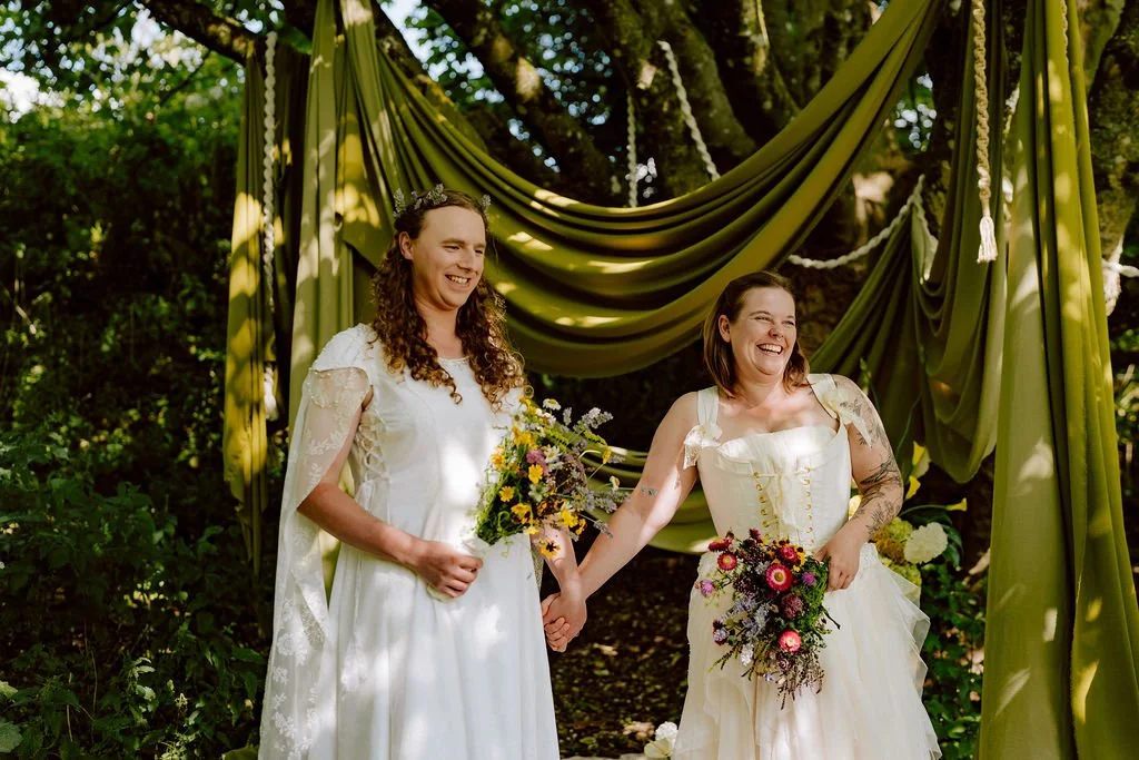 two brides under a tree canopy holding home made bouquets and smiling