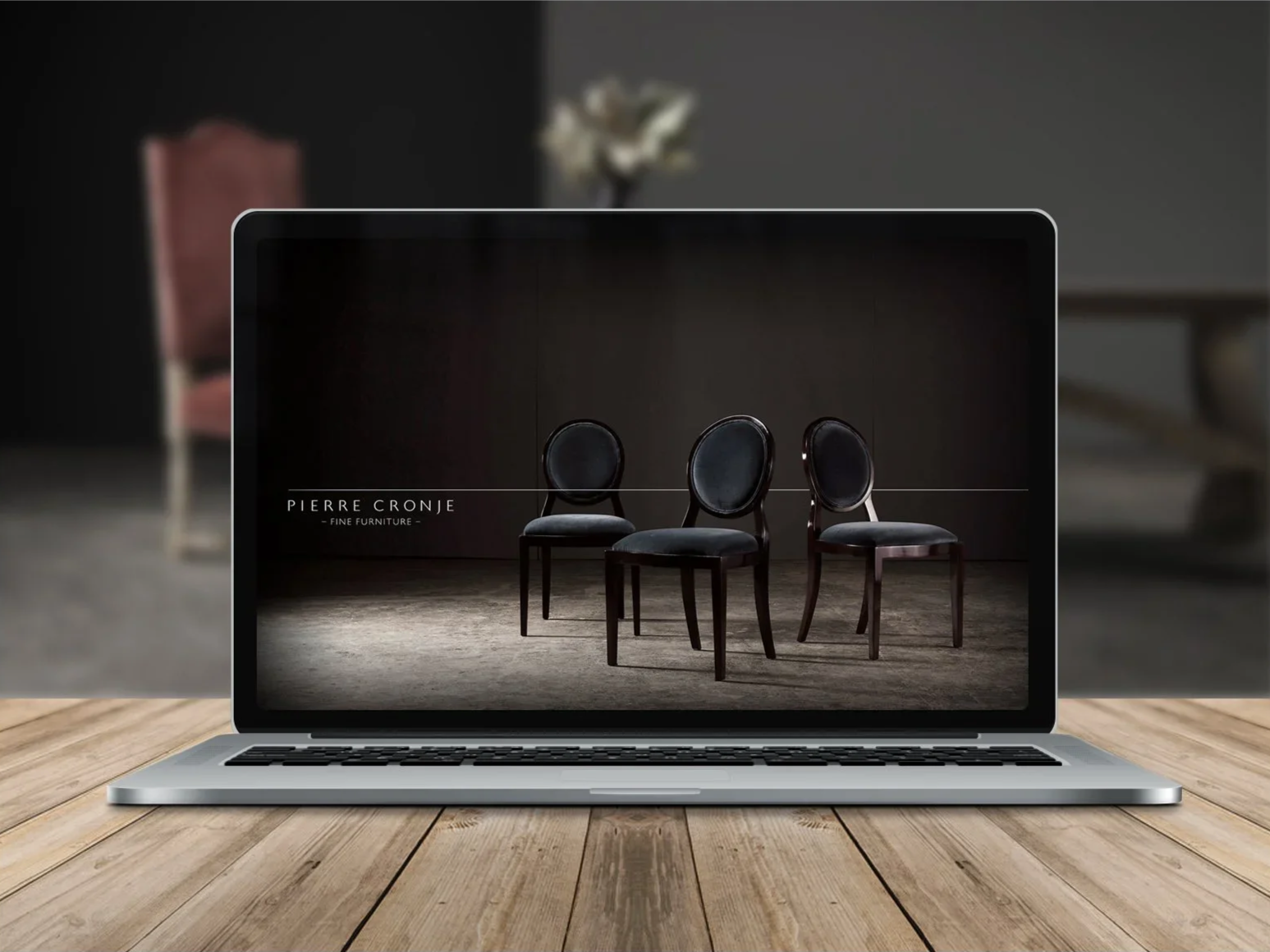 Laptop displaying an image of three dark-colored chairs with rounded backs in a row on a wooden floor, with text that reads 'Pierre Cronje Fine Furniture'.