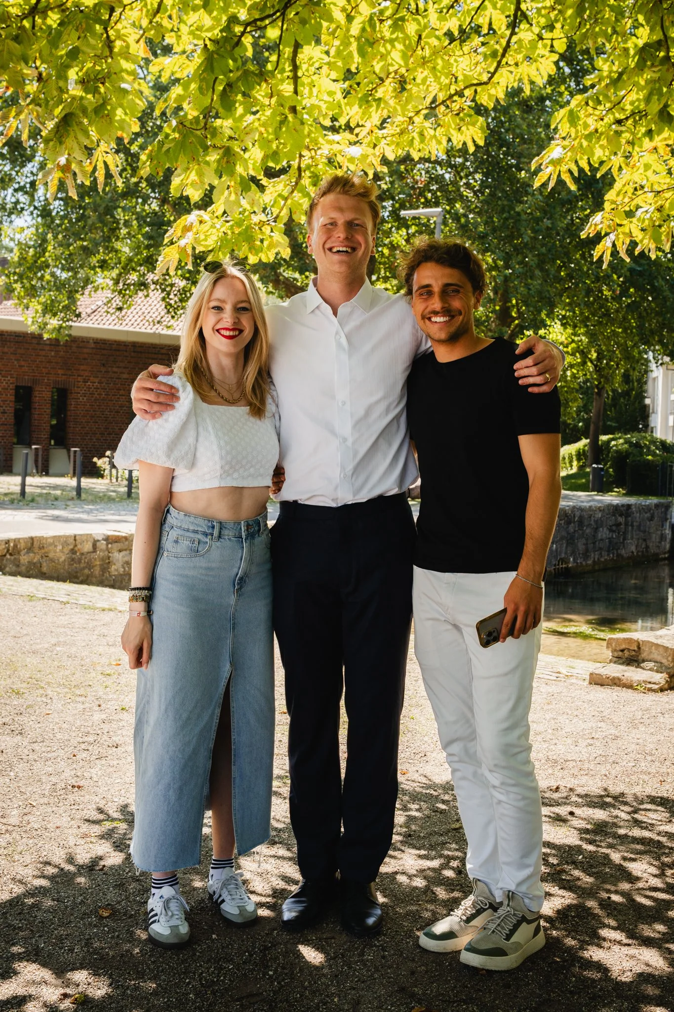 Drei junge Erwachsene, zwei Männer und eine Frau, stehen unter einem Baum im Freien und lachen. Sie tragen sommerliche Kleidung, die Frau hat lange blonde Haare, der Mann in der Mitte hat blondes Haar, der andere Mann hat dunkle, lockige Haare.