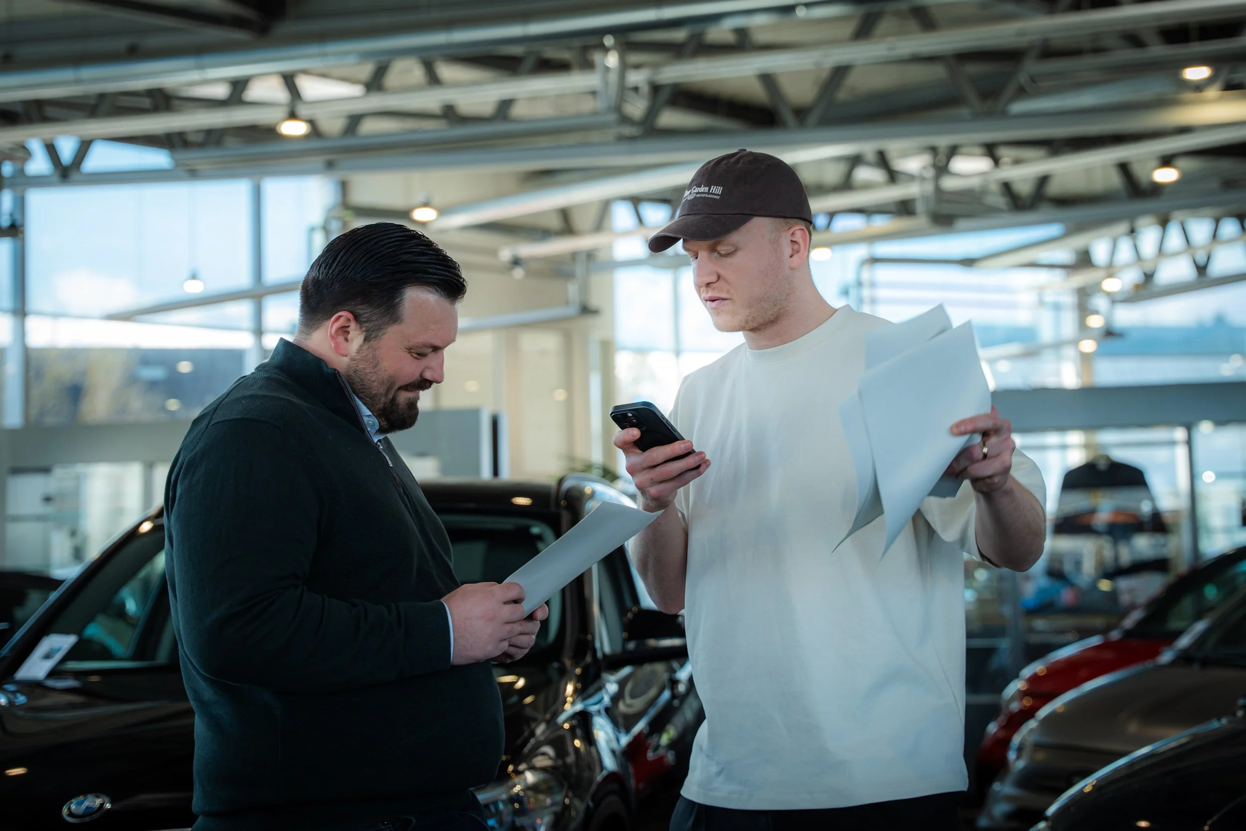 Zwei Männer in einem Autohaus, die sich miteinander unterhalten und Dokumente sowie ein Smartphone in den Händen halten.