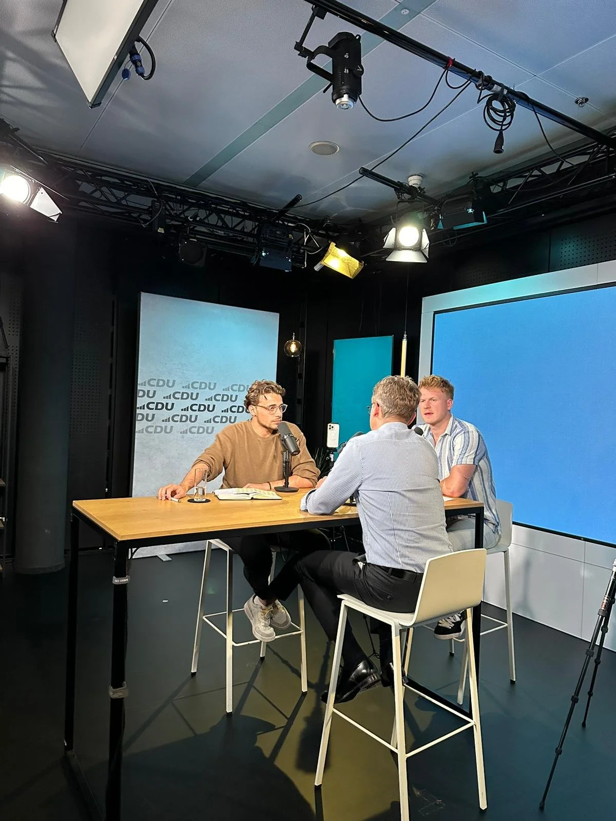 Drei Männer diskutieren während eines Podcasts oder einer Radioaufnahme im Studio, mit einem Tisch, einem Mikrofon, einem großen Bildschirm im Hintergrund und professioneller Beleuchtung