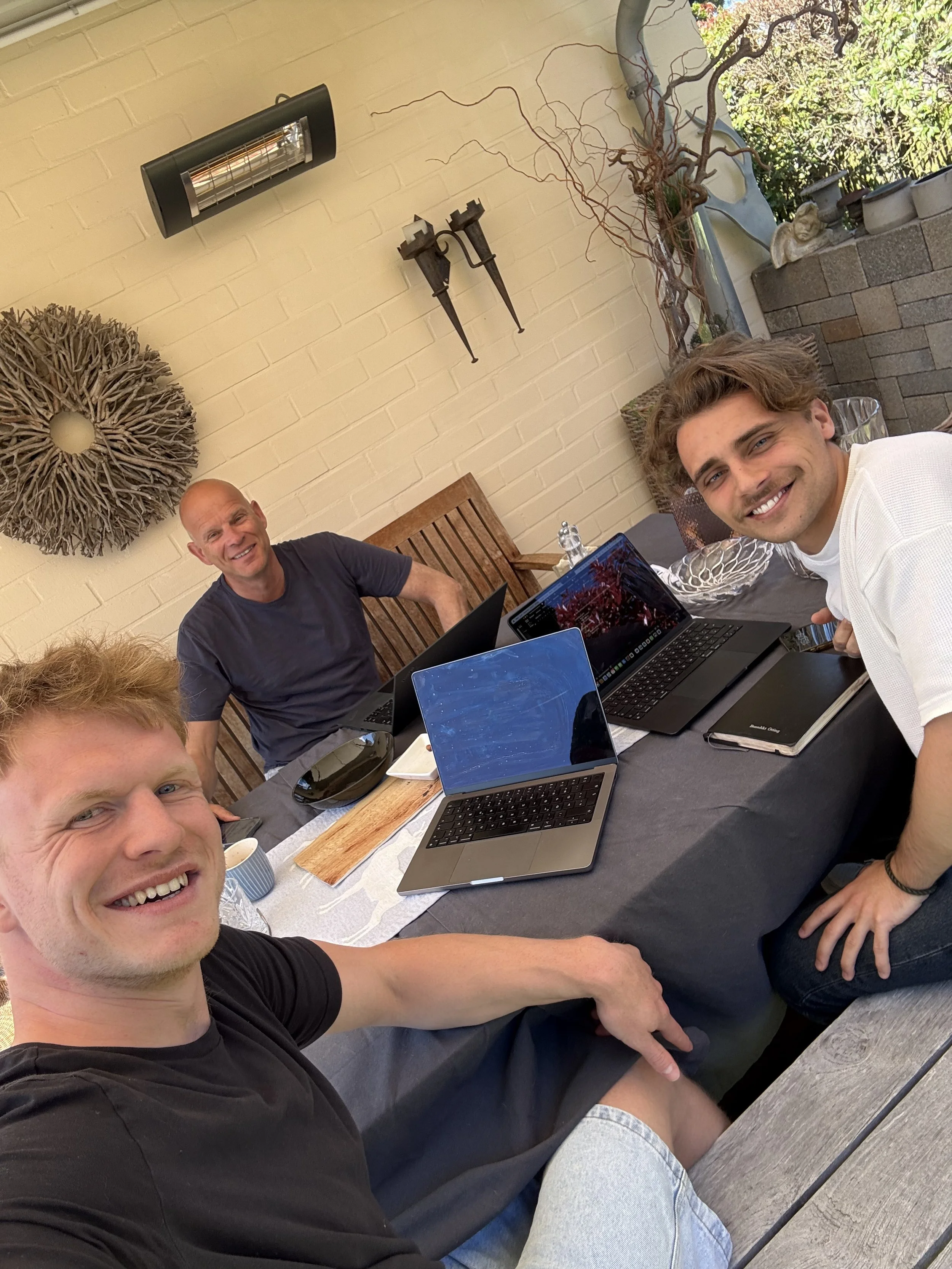 Vier Männer sitzen bei einem Treffen auf einer Terrasse mit Laptops. Es sind drei jüngere Männer und ein älterer Mann. Im Hintergrund befinden sich Dekorationsgegenstände an einer hellen Wand.