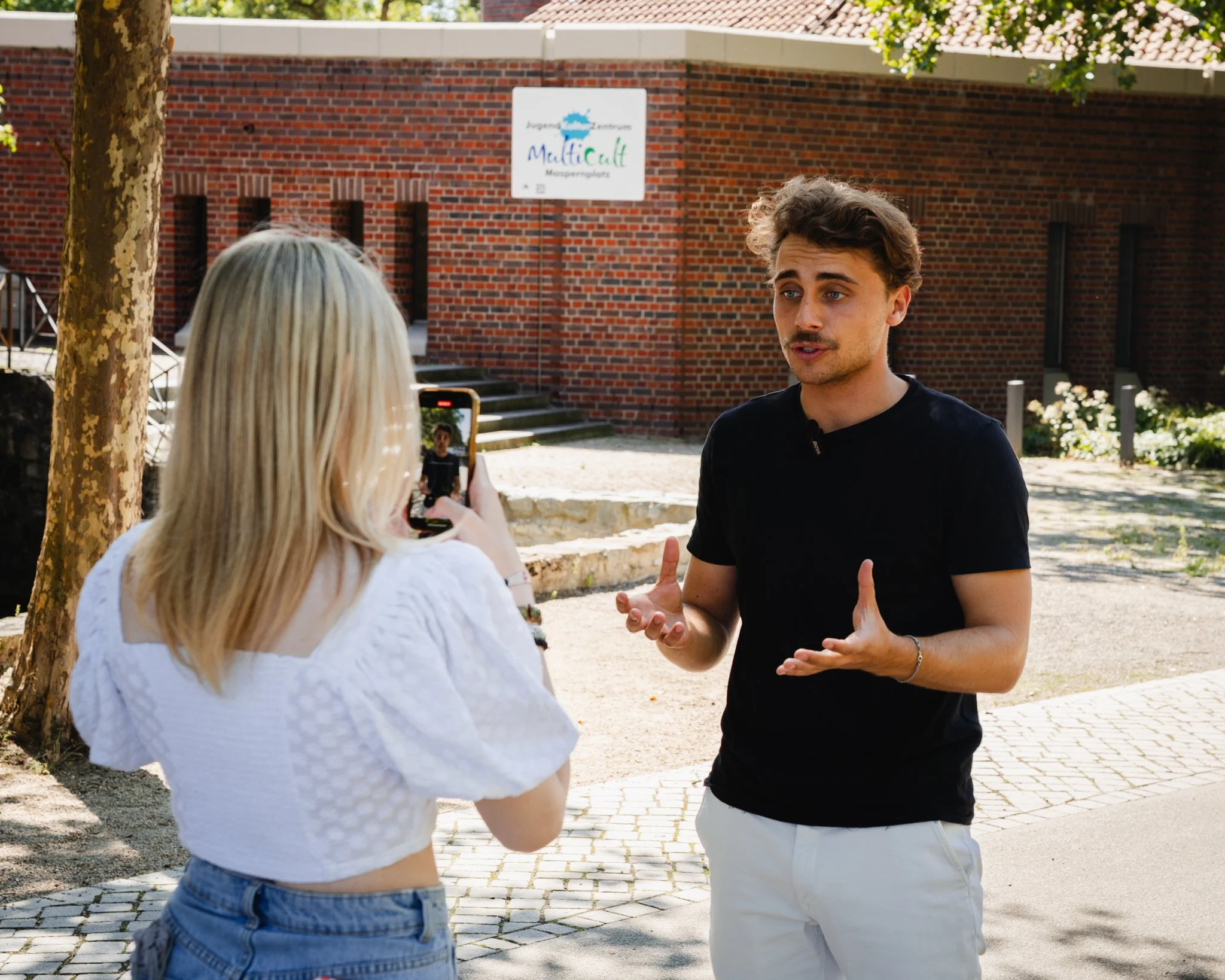 Ein junger Mann spricht während eines Interviews mit einer Frau, die ein Smartphone zum Aufnehmen verwendet, im Freien vor einem roten Backsteingebäude mit einem Baum auf der linken Seite.