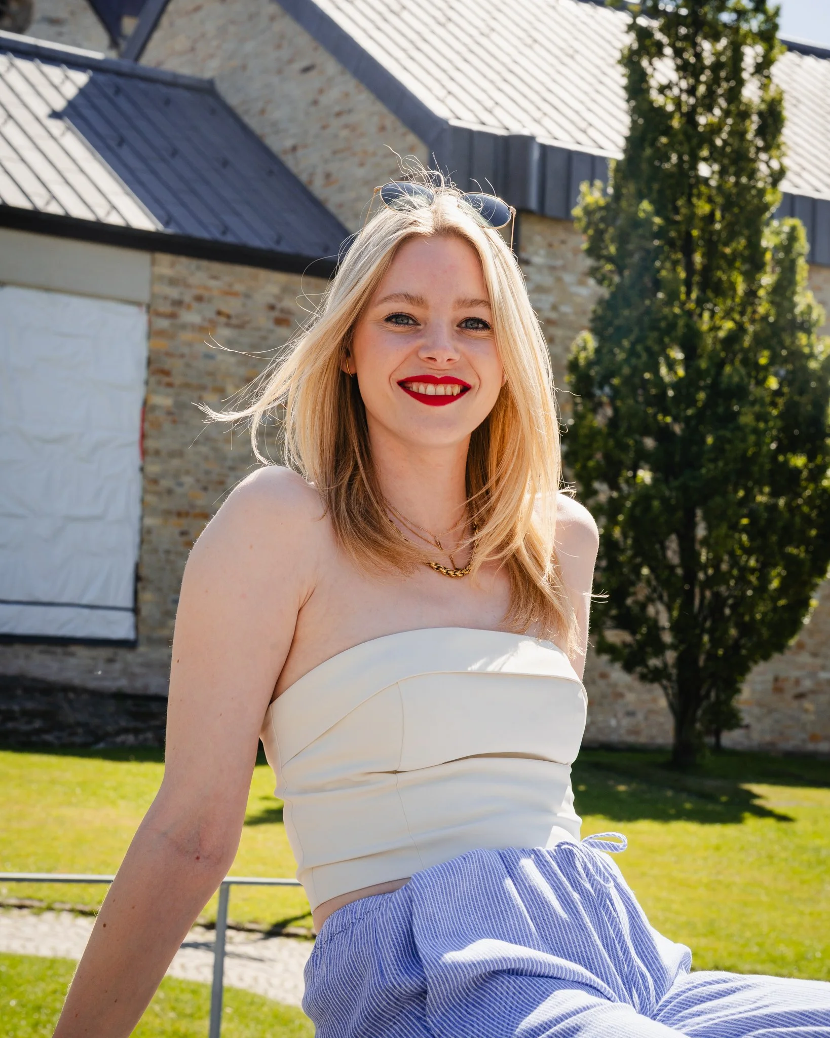Junge Frau mit roten Lippen, Sonnenbrille auf dem Kopf, lächelnd im Freien bei Sonnenschein, trägt eine weiße Schulterfrei-Top und blaue Hosen, im Hintergrund ein Baum und ein altes Gebäude.