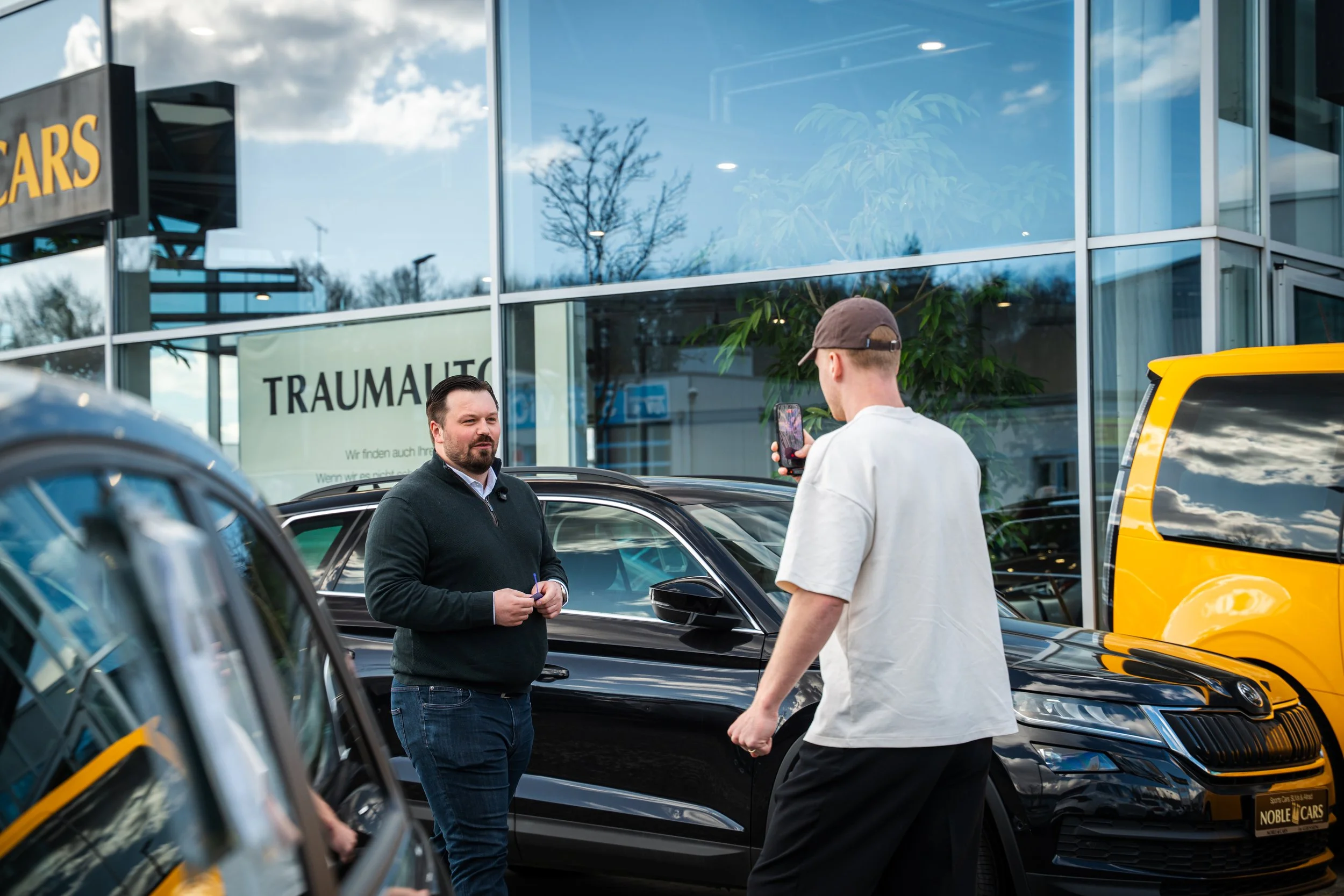 Zwei Männer stehen vor einem schwarzen Auto und unterhalten sich, während ein Dritter ein Foto mit einem Handy macht, vor einem Autohaus mit Glasfassade.