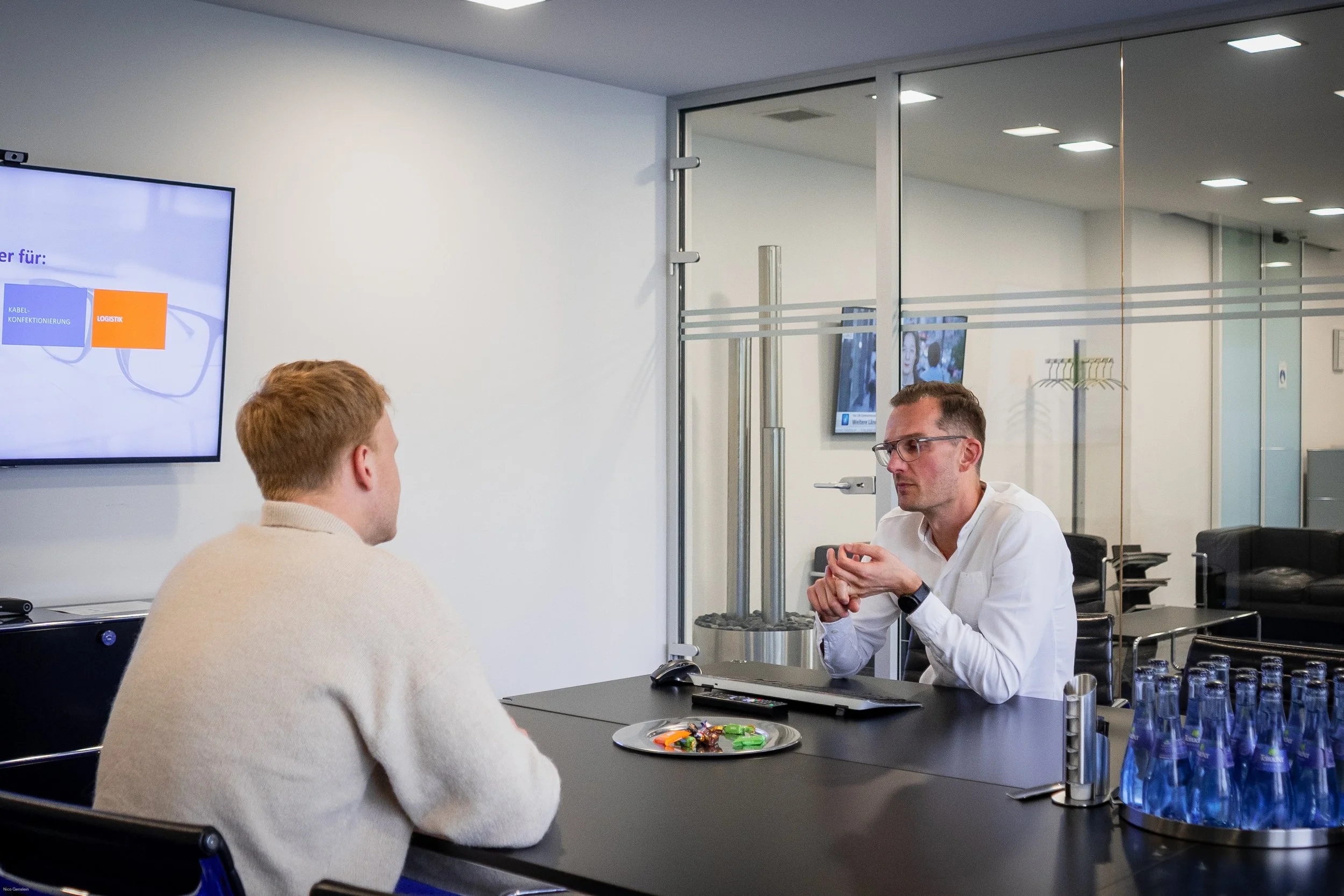 Zwei Männer sitzen in einem Büro an einem Tisch im Gespräch. Auf dem Tisch liegt ein Laptop, ein Tablett mit Süßigkeiten, eine Tasse, und einen Container mit Wasserflaschen. Im Hintergrund sind Monitore und ein Besprechungsraum sichtbar.