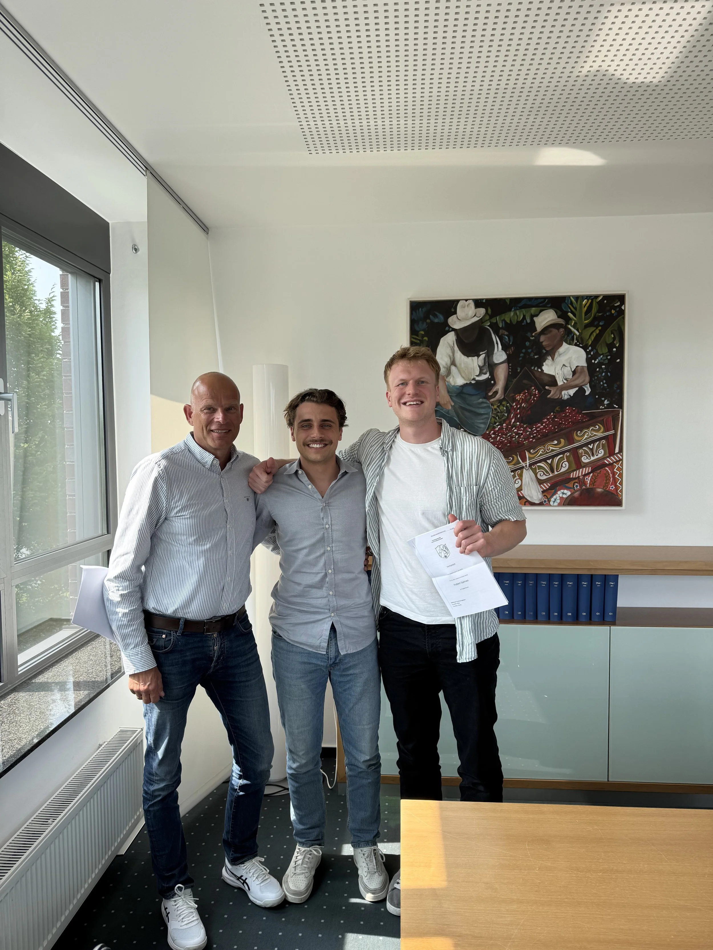 Drei junge Männer stehen lächelnd in einem modernen Büro, eines hält ein Dokument. Im Hintergrund ist ein kunstvolles Gemälde, eine Fensterseite mit Blick nach draußen, und eine Bücherecke zu sehen.