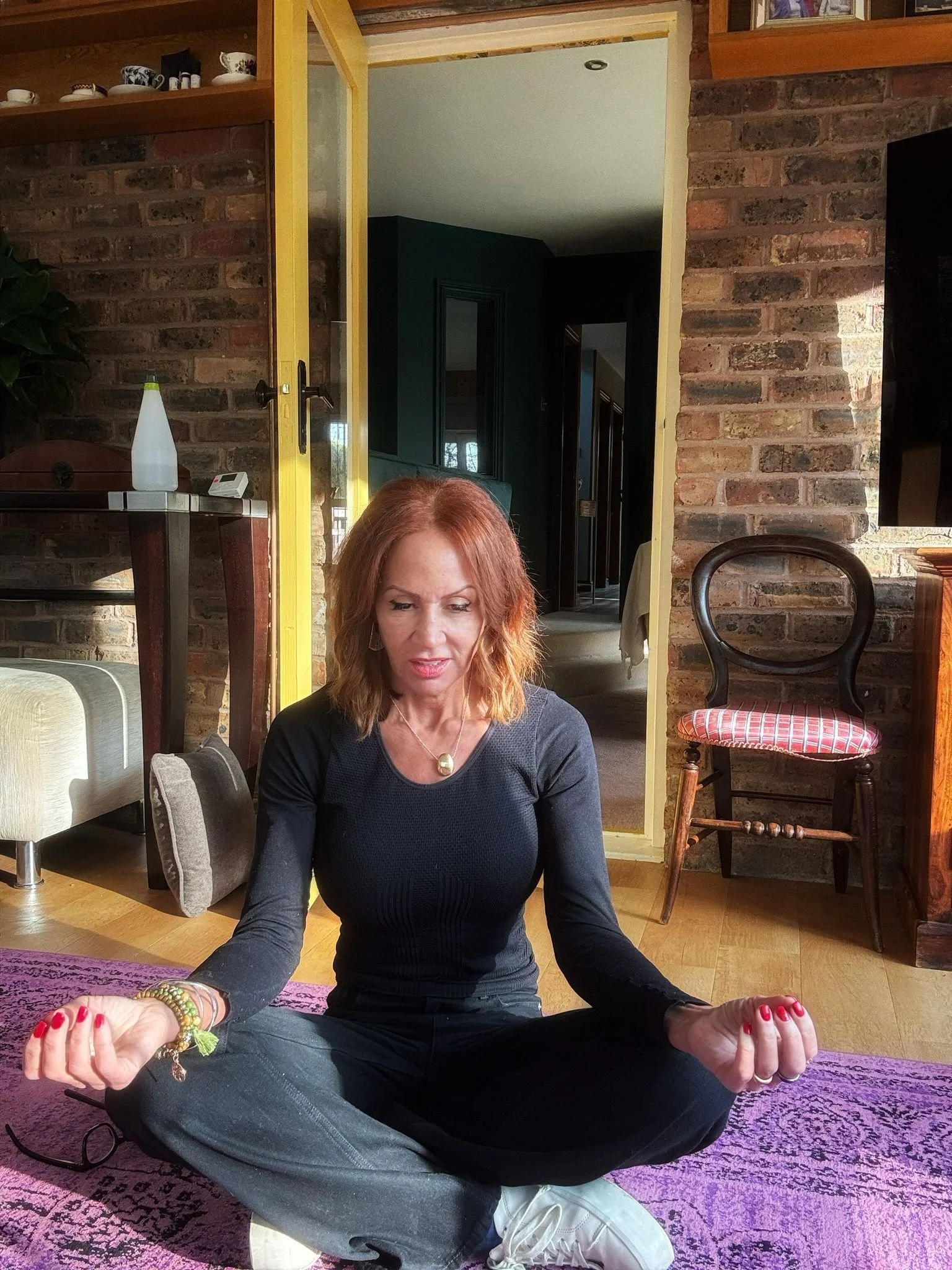 A woman with red hair, dressed in black, sitting cross-legged on a pink rug, practicing meditation indoors.