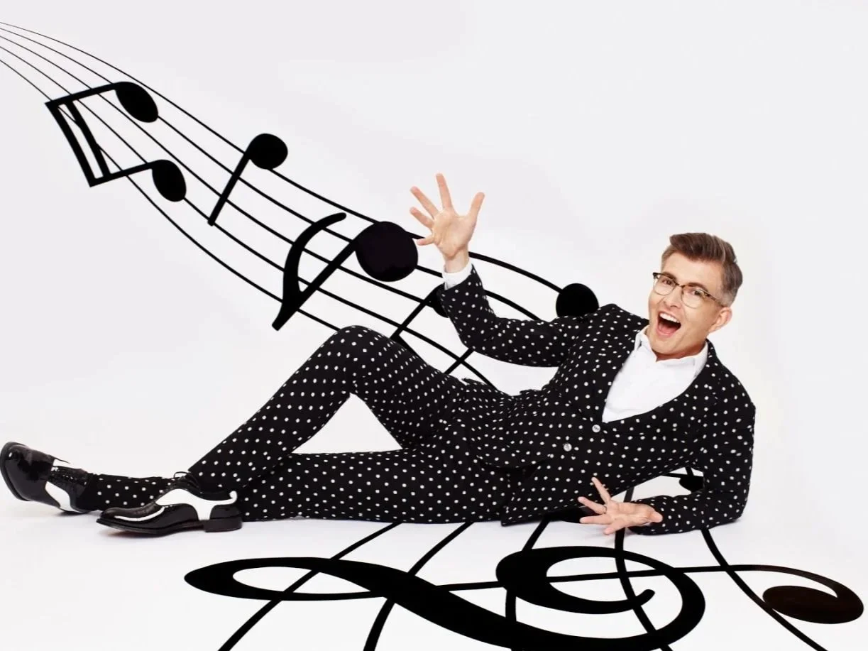 A man in a polka dot suit with glasses lying on the floor, posing happily, with black musical notes and staff lines illustrated on the white background around him.