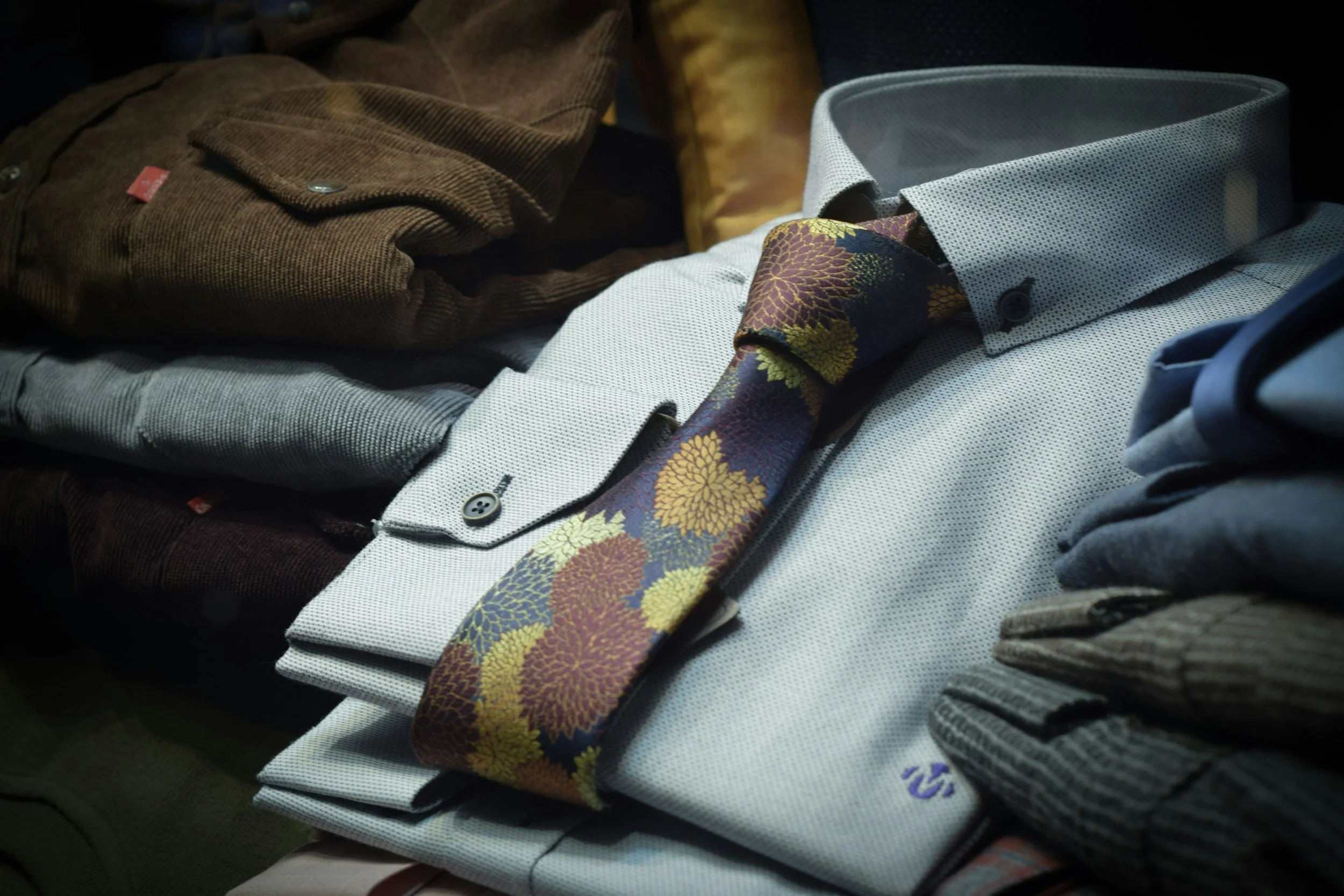A pile of folded men's dress shirts including a brown corduroy shirt, a white shirt with a patterned tie, and other shirts in various colors and textures.