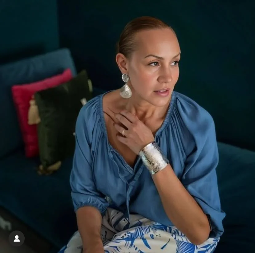 A woman with light skin and brown hair styled slicked back, wearing large earrings and a blue blouse, sits on a dark sofa with colorful pillows in the background, looking to the side with her hand on her neck.