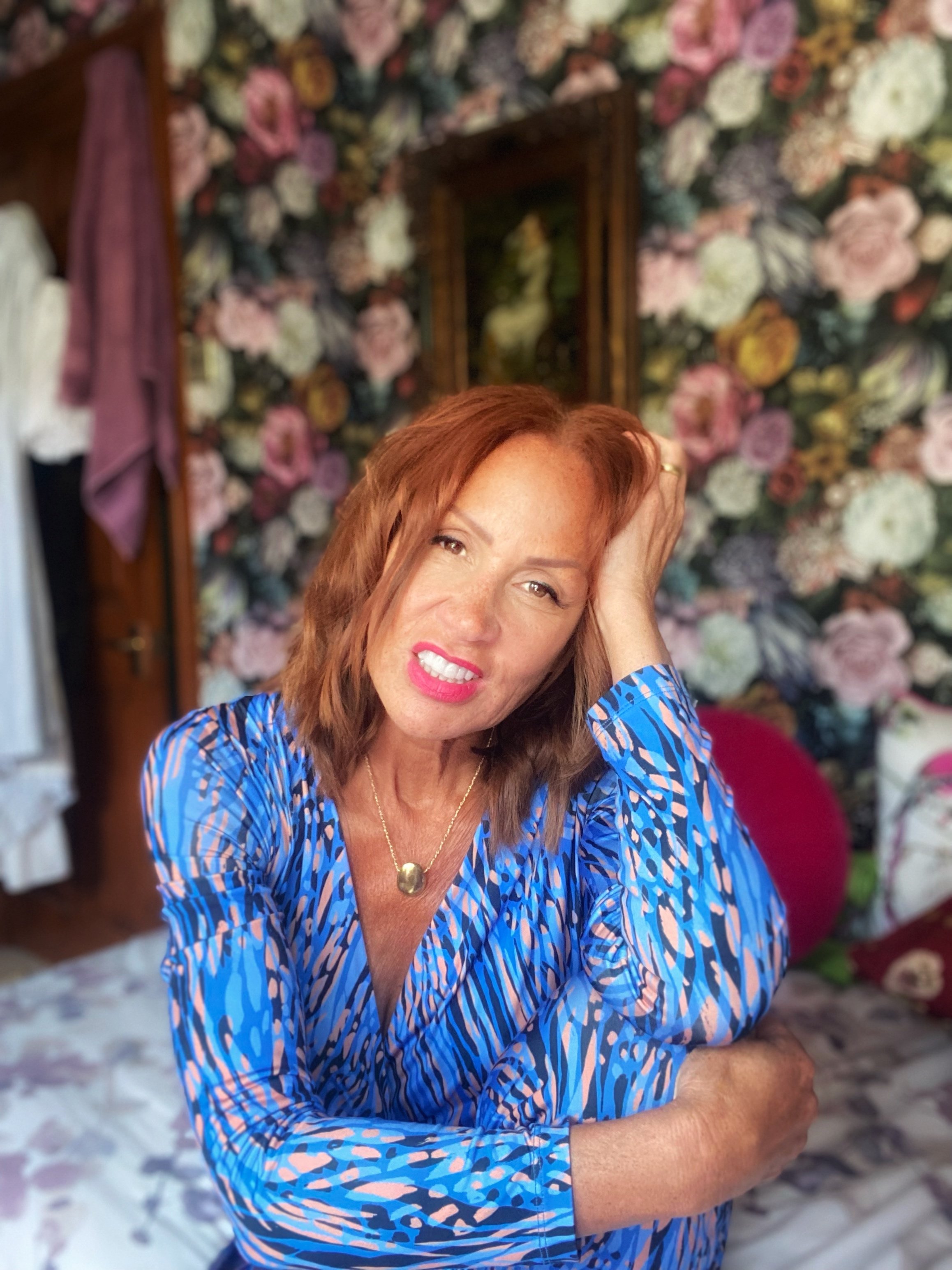 A woman with red hair, wearing a blue patterned shirt and a gold necklace, sitting in a floral decorated room with a distressed expression, resting her head on her hand.