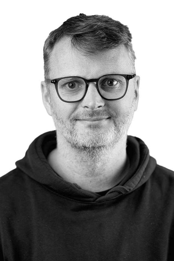 En mann med brunt hår og skjegg, iført svarte briller