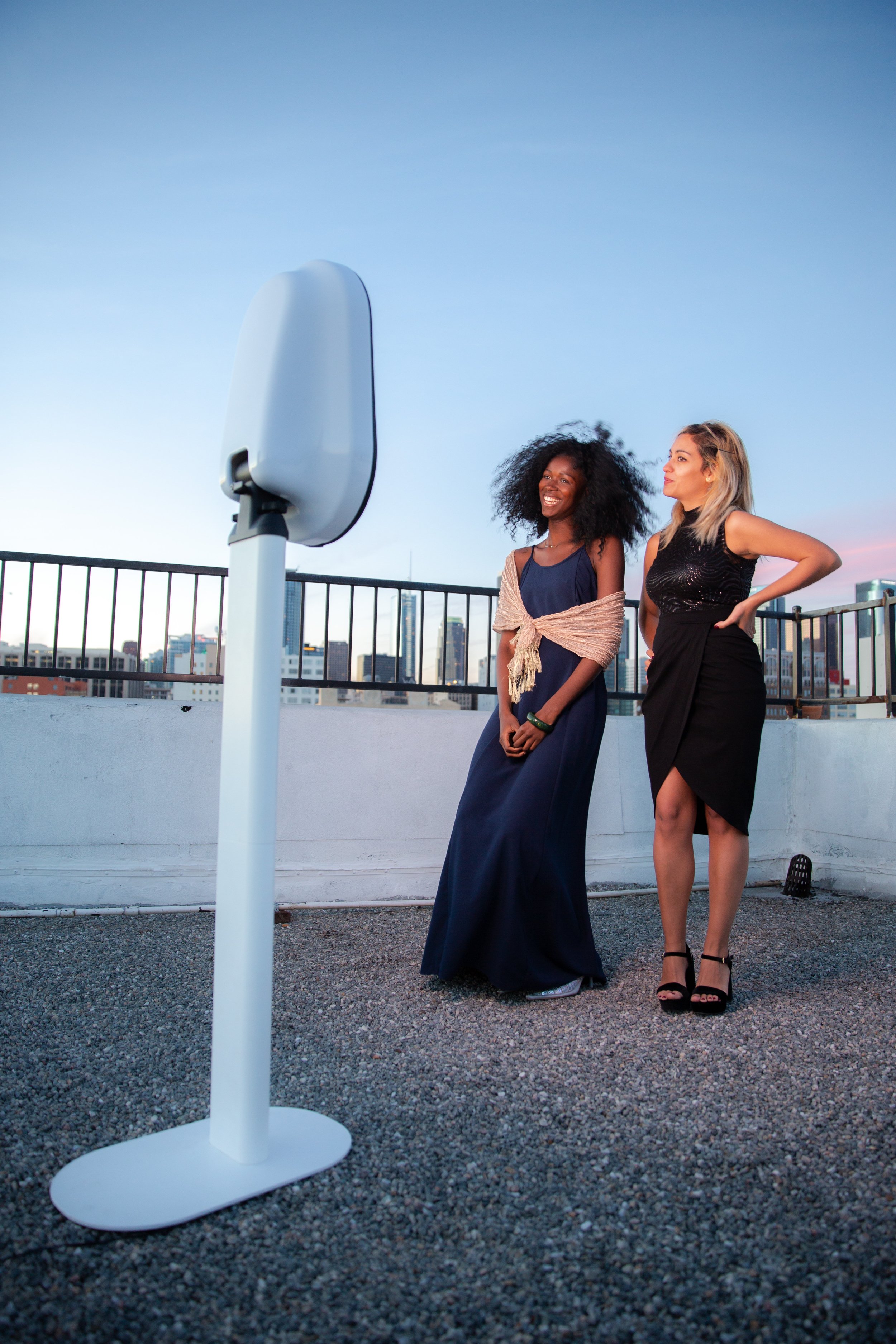 Two women standing outdoors on a rooftop, next to a modern photo booth, with city skyline in the background.
