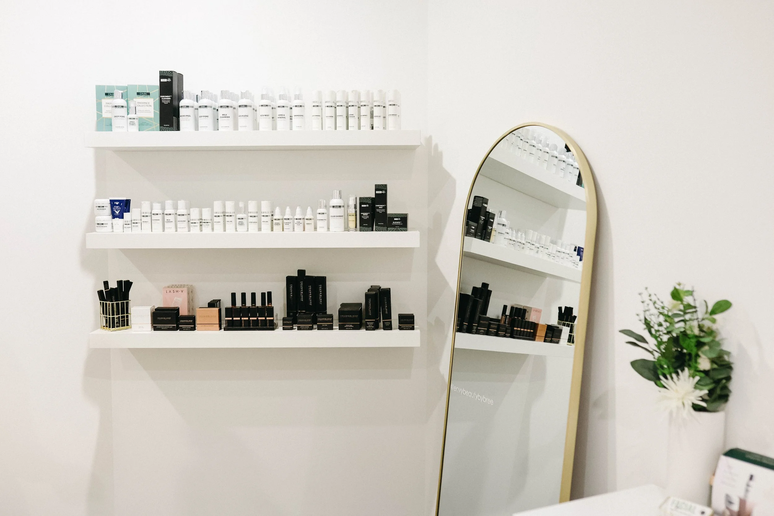Display of skincare and beauty products on white shelves, a gold-framed mirror, and a potted plant in a clean, minimalist room