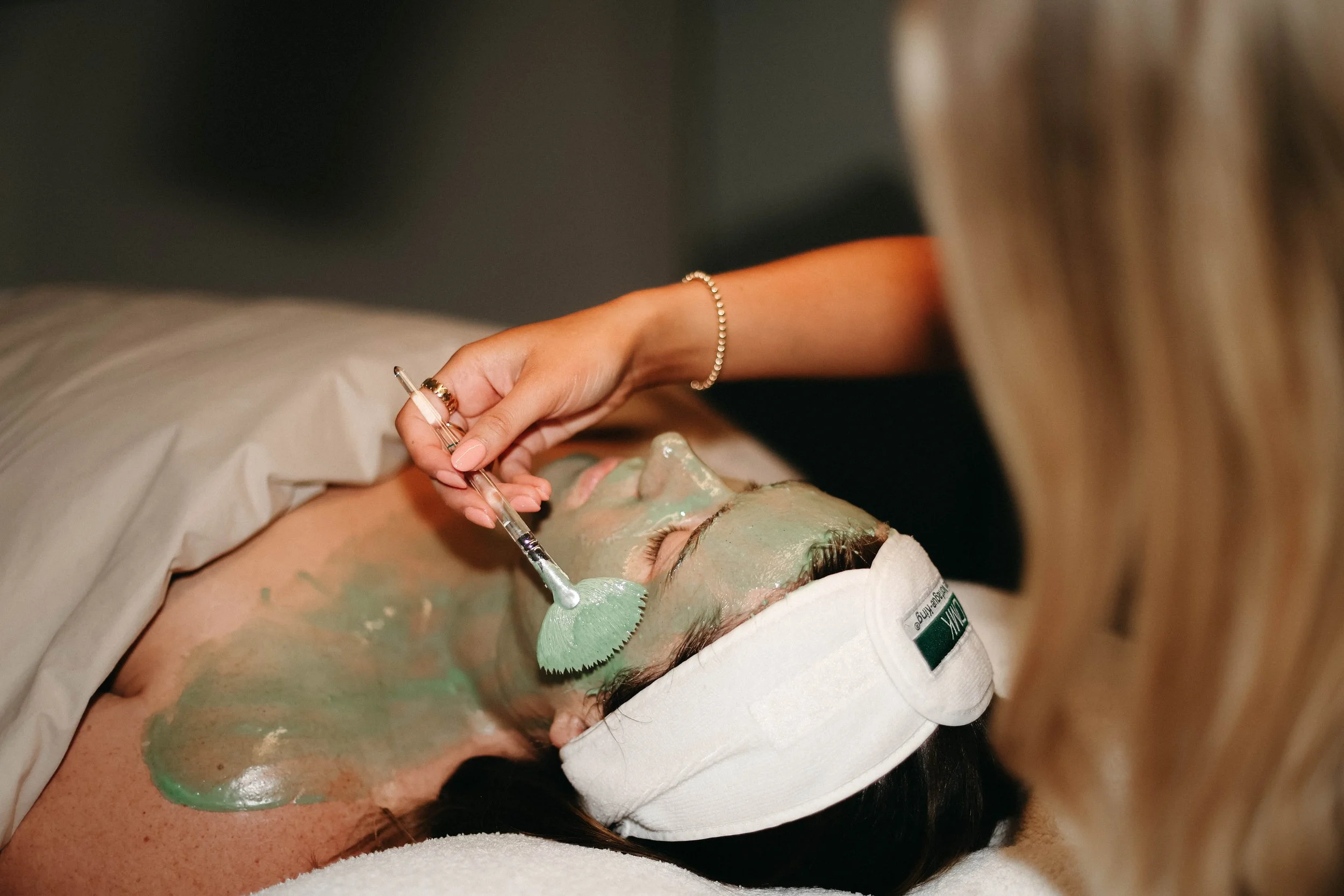 Person receiving a facial treatment with a green facial mask applied, lying on a spa bed with a towel and a headband.