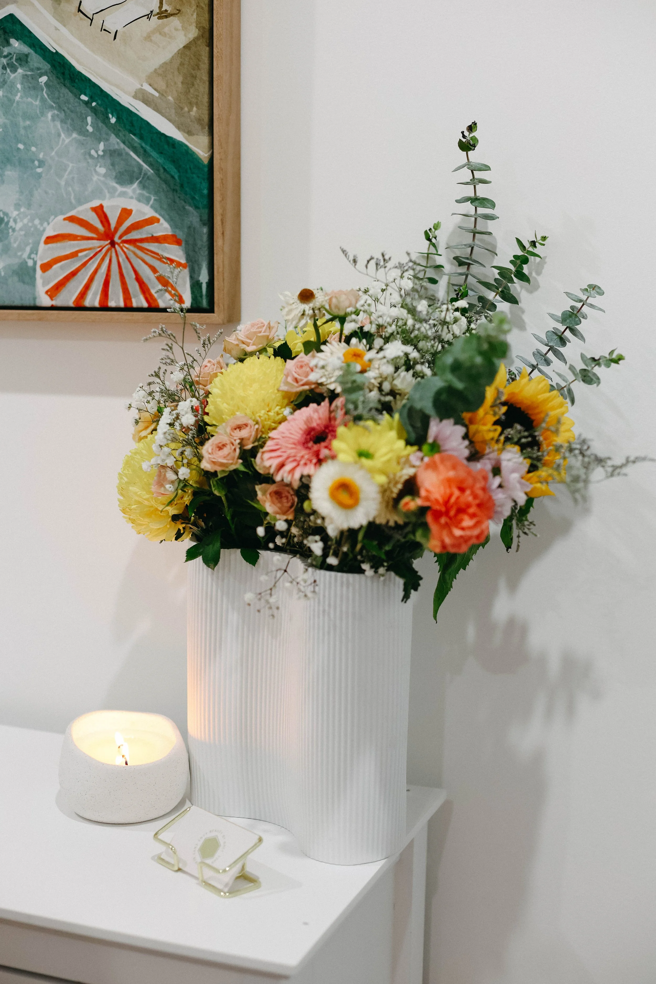 A tall, white, ribbed vase holding a colorful bouquet of flowers including lilies, roses, daisies, and eucalyptus leaves, with a small lit candle and a decorative paperclip on a white table against a white wall with a framed abstract artwork.