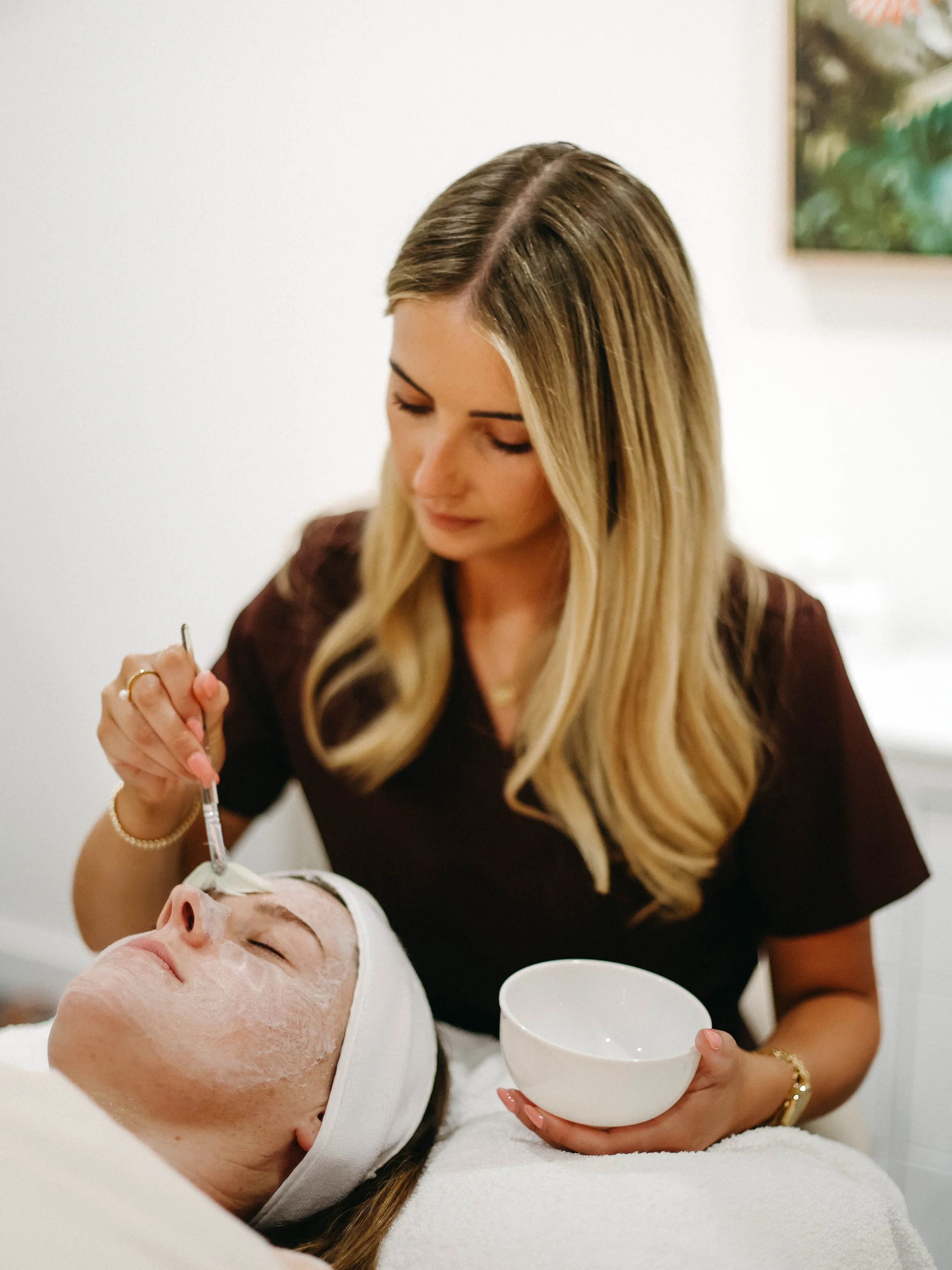 A woman with long blonde hair in a brown top is administering a facial treatment to another woman lying down with a white headband, closed eyes, and a white face mask, in a spa setting.