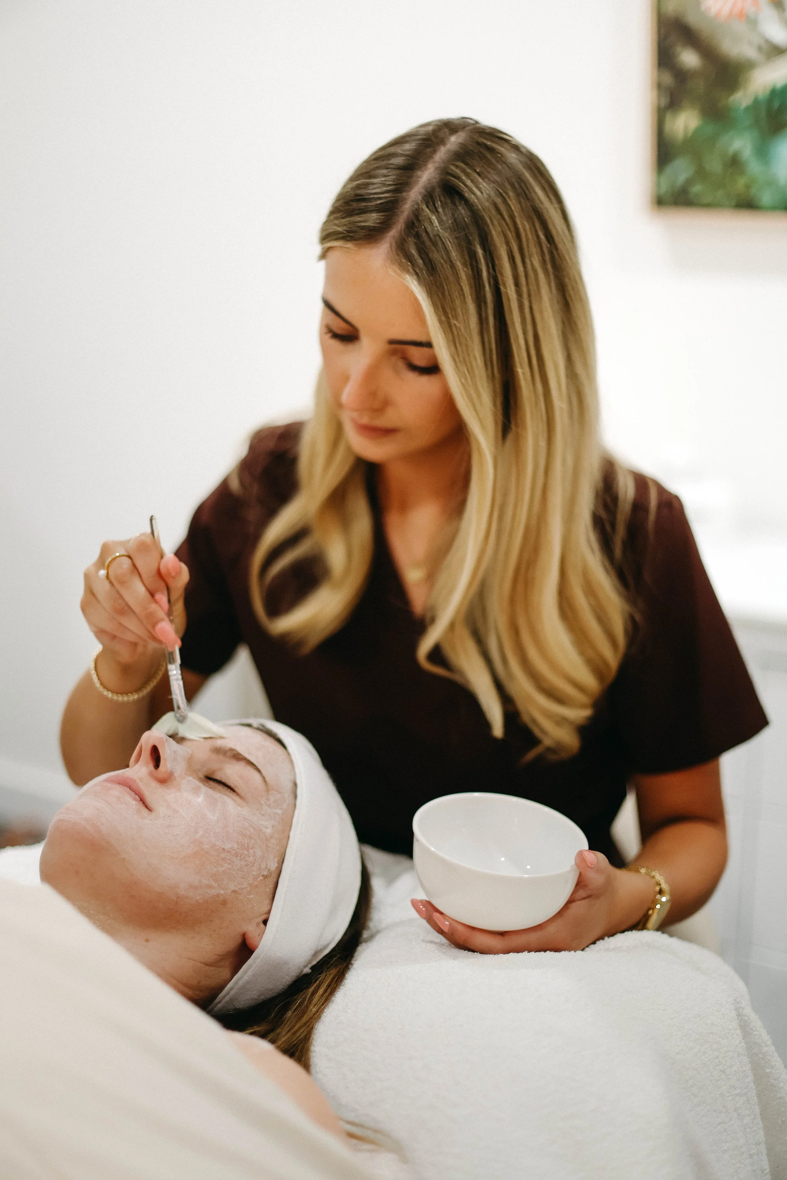 A woman in a brown top is applying a facial treatment to a woman lying on a massage table, covered with a white towel. The woman receiving the treatment has a headband and her eyes closed.