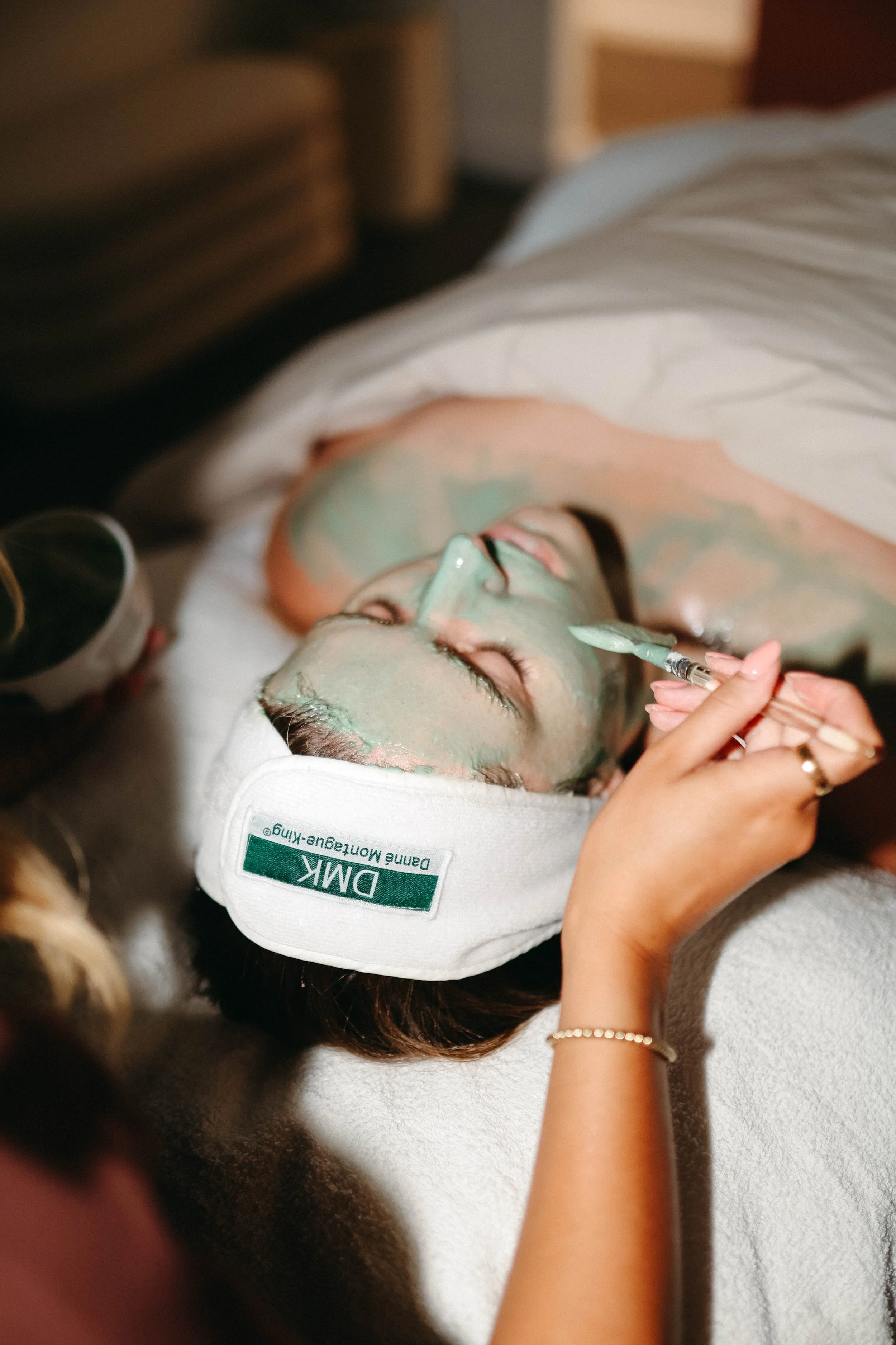 A woman lying down with her eyes closed, receiving a facial treatment with a green mask, wearing a white headband labeled 'DMK' and 'Danish Manufacturing,' holding a brush in her right hand.