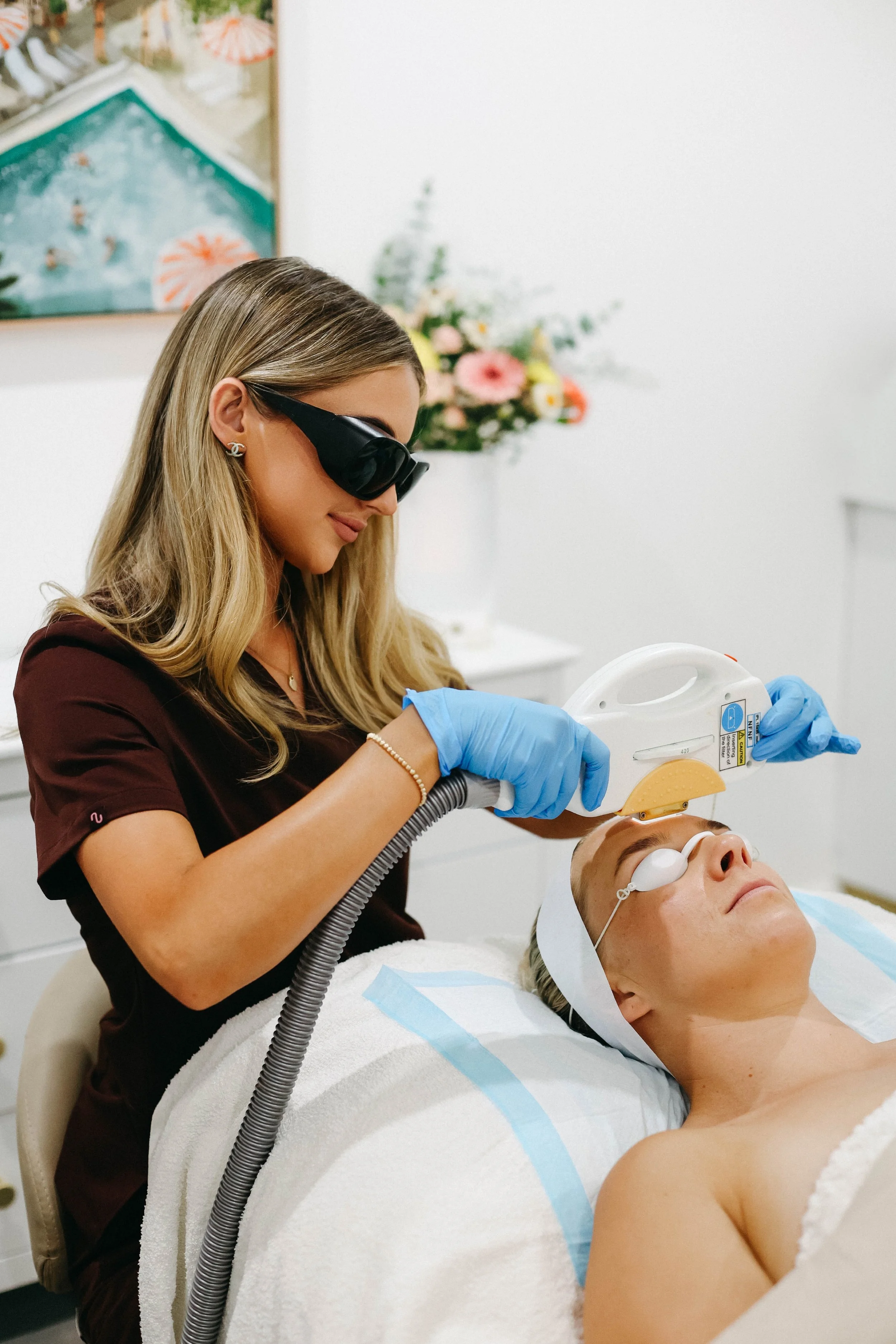 A woman in protective eyewear and gloves performs a cosmetic laser procedure on a patient lying on a treatment bed in a clinic.