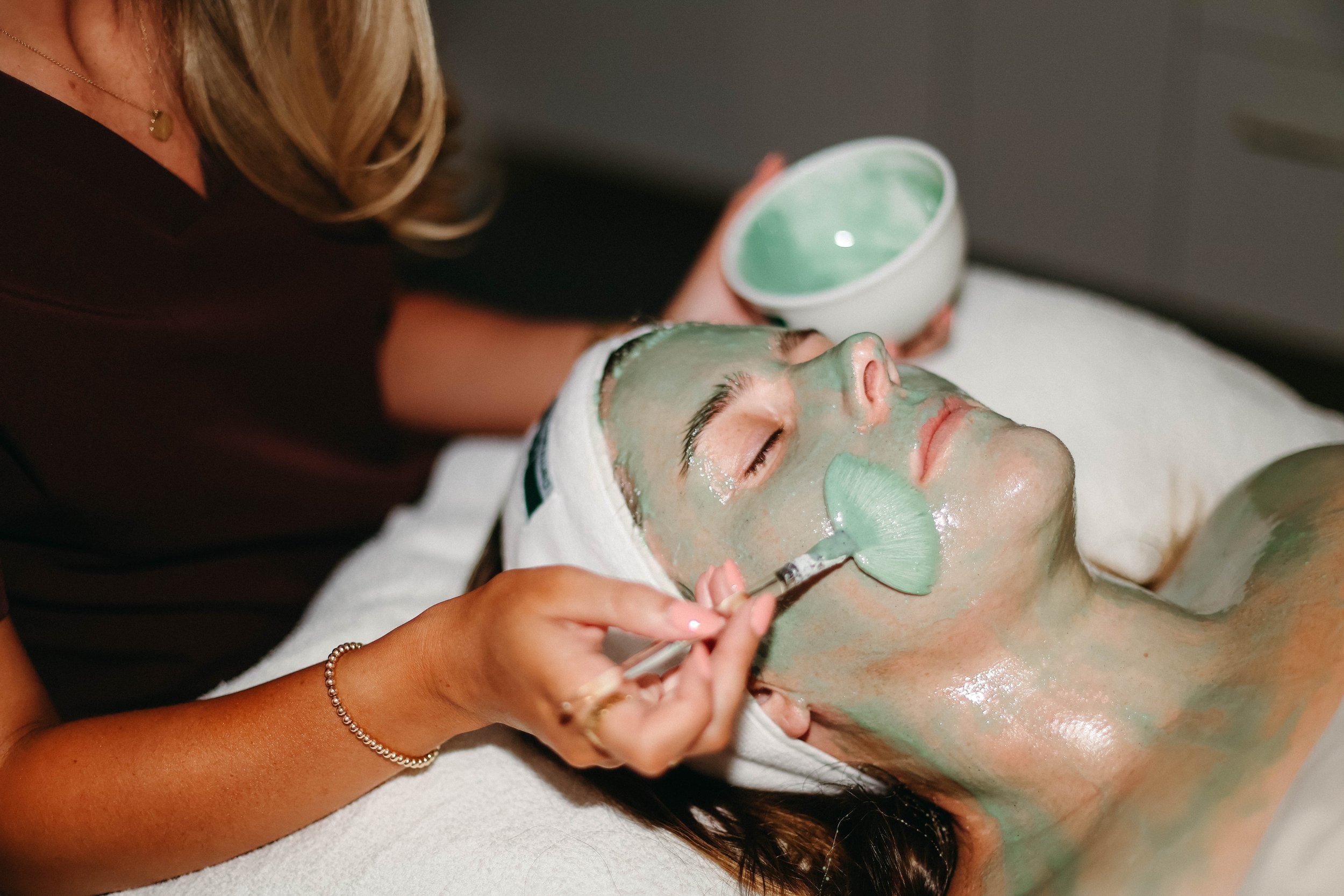 A woman receives a facial treatment with a green mask applied to her face by an esthetician in a spa setting.
