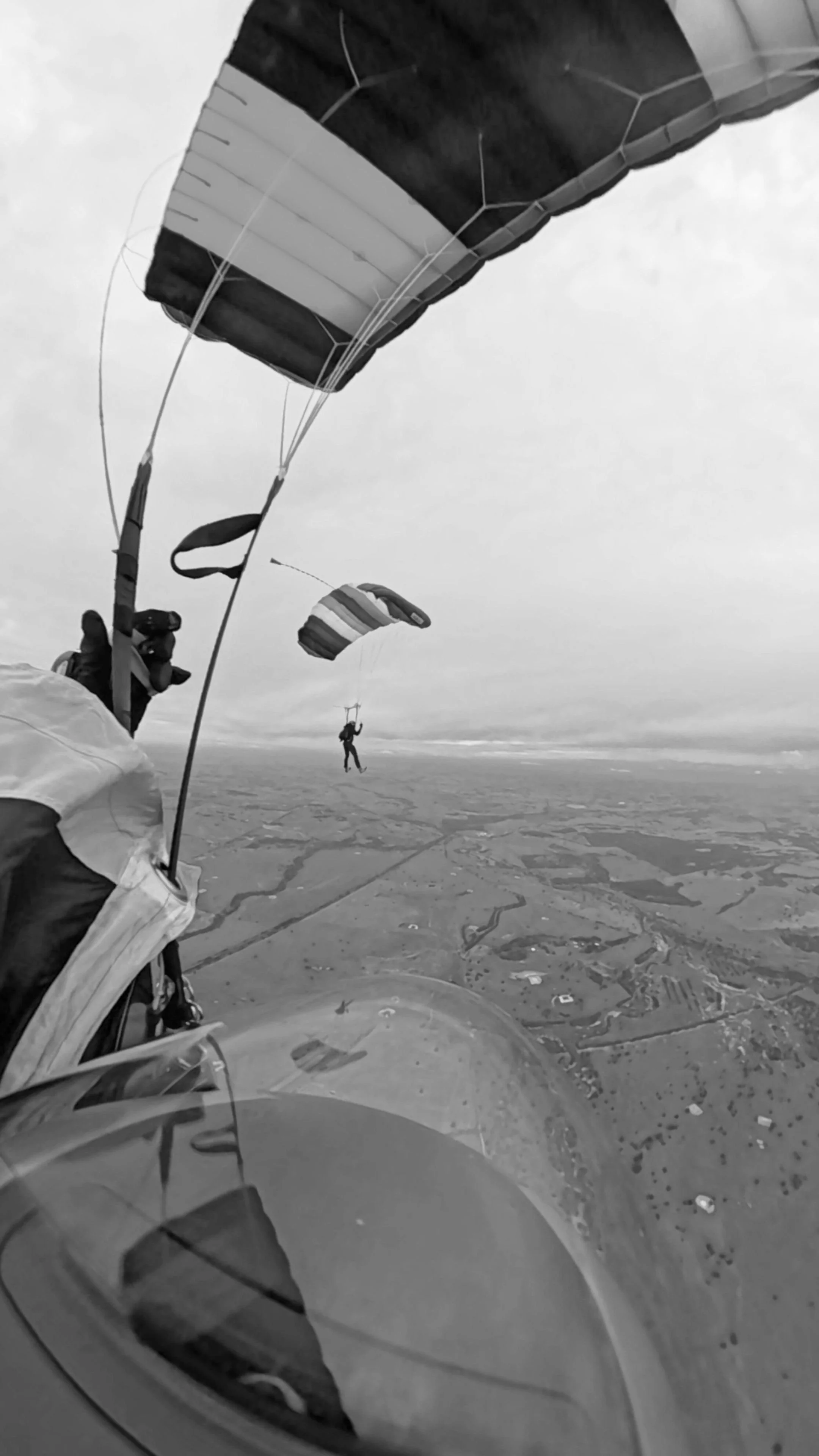 Two parachuters are gliding in the sky over a landscape, with one parachute fully open in the foreground and another, smaller parachute in the background.