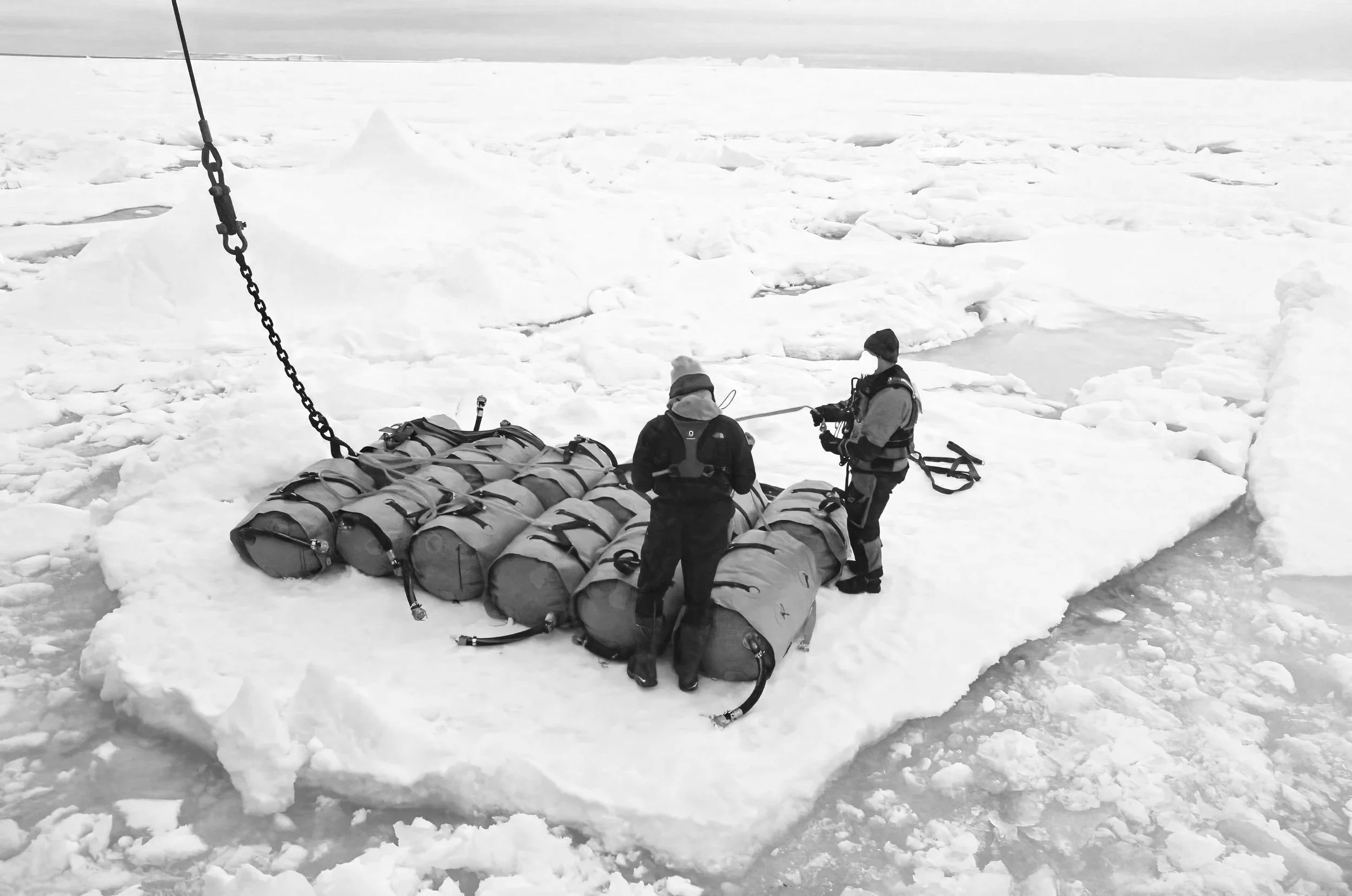 Two people in winter clothing stand on snow and ice, surrounded by a large pile of waterproof gear or bags, with a boat and frozen water in the background.