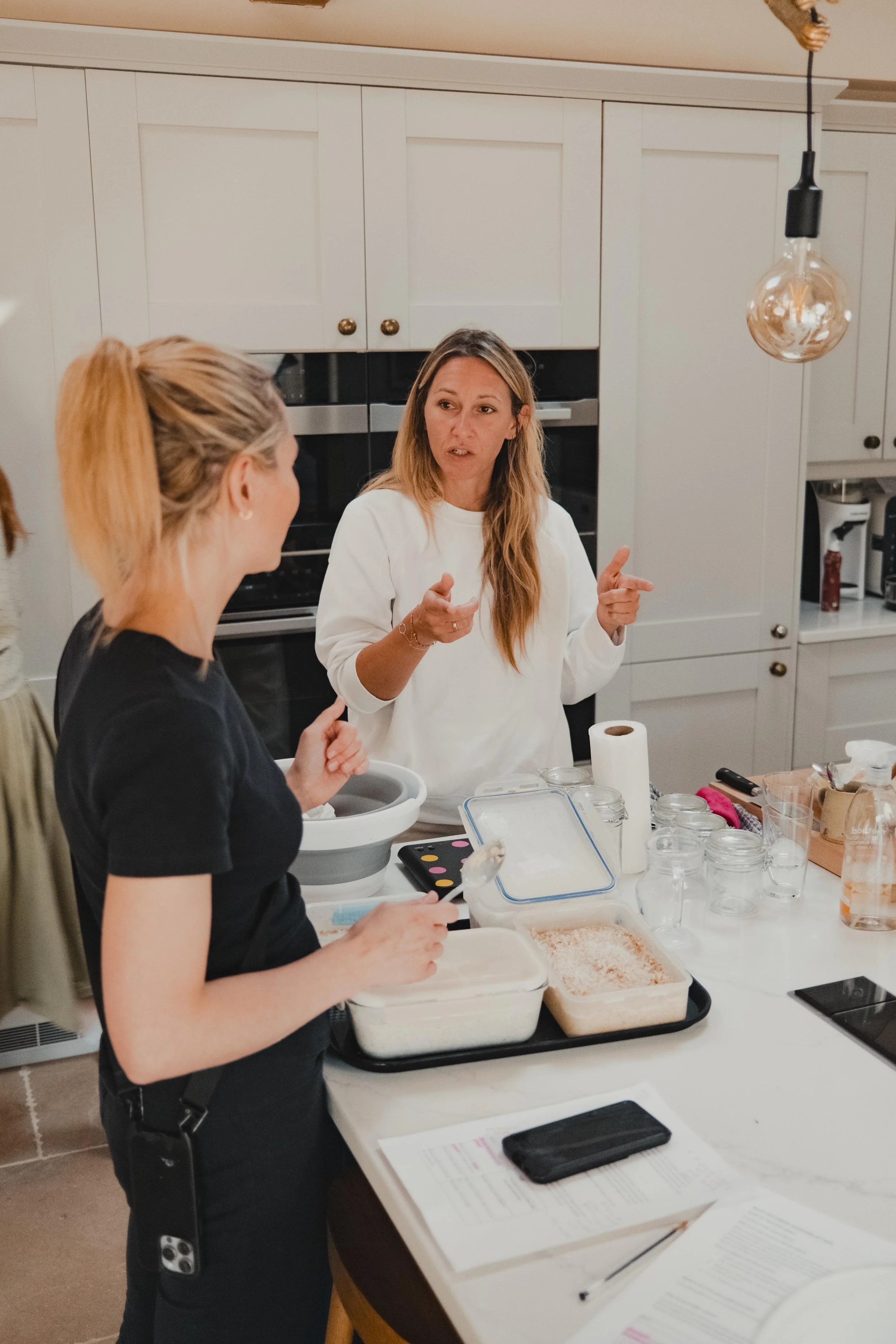 Two women having a discussion in a kitchen, one holding a spoon and the other gesturing with her hands, surrounded by baking ingredients and utensils on the counter.